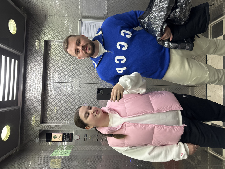 Two people are in an elevator, taking a selfie. The woman has a pink vest and her hair is in a braid. The man wears a blue sweater with "CCCP" in white letters and holds a dark jacket. The elevator interior is metallic with geometric patterns and light fixtures, featuring a control panel with buttons and directional arrows.