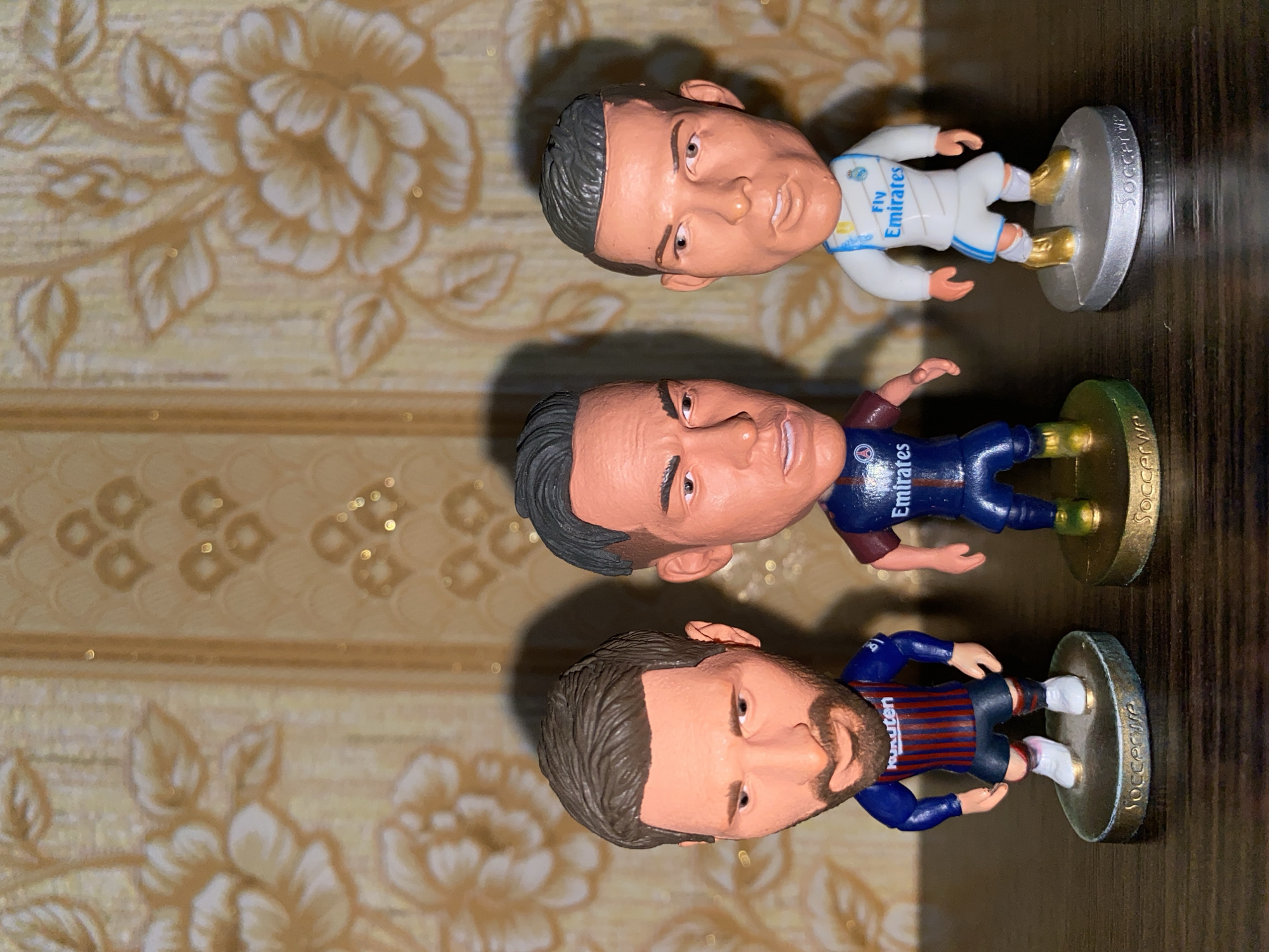 The image shows three bobblehead figurines of soccer players. From top to bottom, the figures appear to be Cristiano Ronaldo in a Real Madrid jersey, Neymar in a Paris Saint-Germain jersey, and Lionel Messi in a Barcelona jersey. The figurines are standing on circular bases labeled "Soccerwe". The background consists of a floral pattern on a surface that transitions into a dark brown surface. The image does not provide any information about the location in Kharkiv, Ukraine.