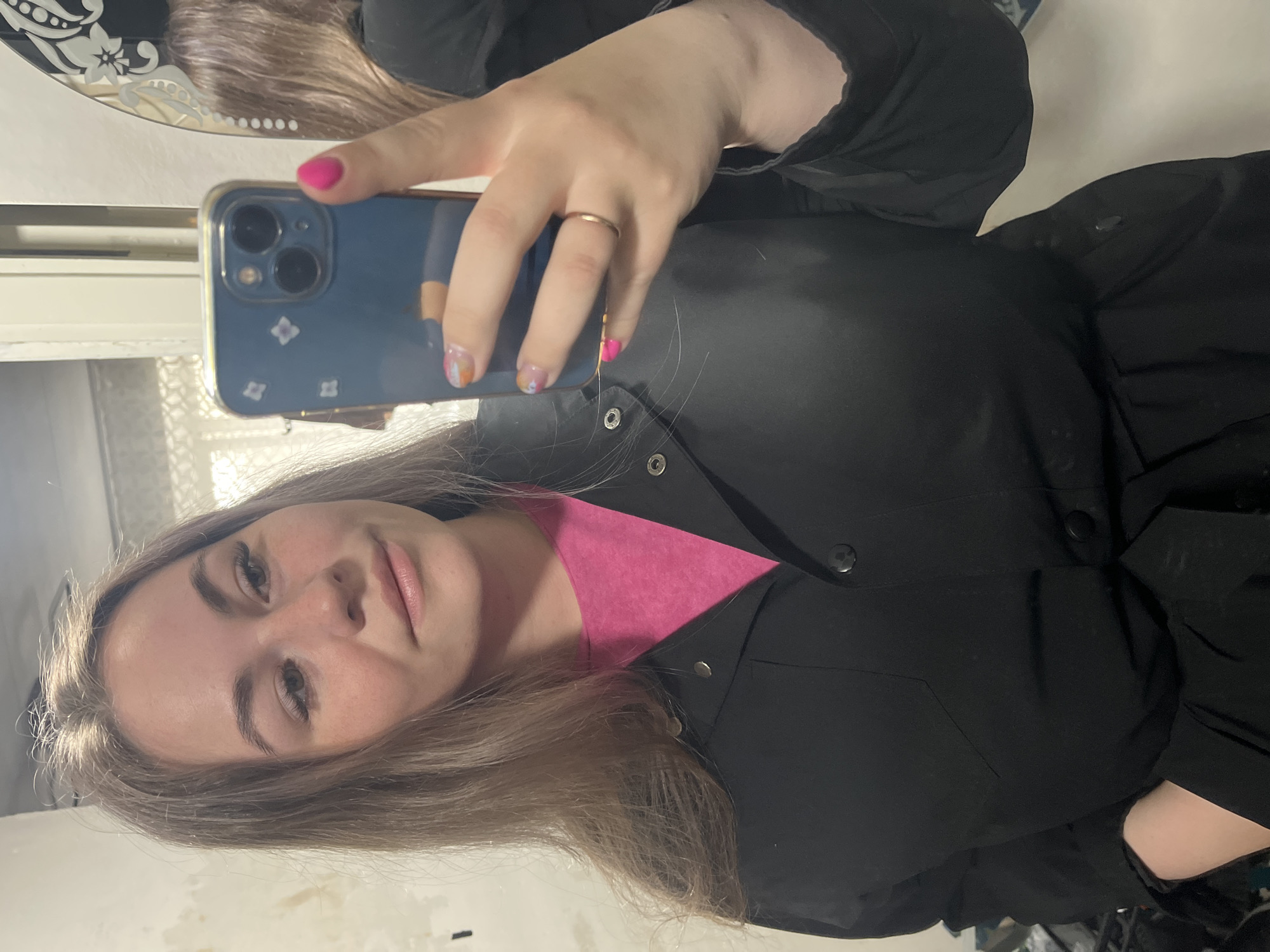 A young woman is taking a selfie in what appears to be an indoor setting. She's wearing a black jacket over a pink top. She is holding a blue smartphone in her right hand with a gold ring on her ring finger. The phone case has a pattern of white flowers or similar shapes. Her nails are painted with a mix of colors, including pink and orange. The location is consistent with an indoor setting, with a mirror visible behind her, reflecting a blurred portion of the room. The background suggests a domestic interior, possibly an apartment, with light-colored walls. There is no indication of the city of Chaikovsky, Russia, or any other specific geographic location.