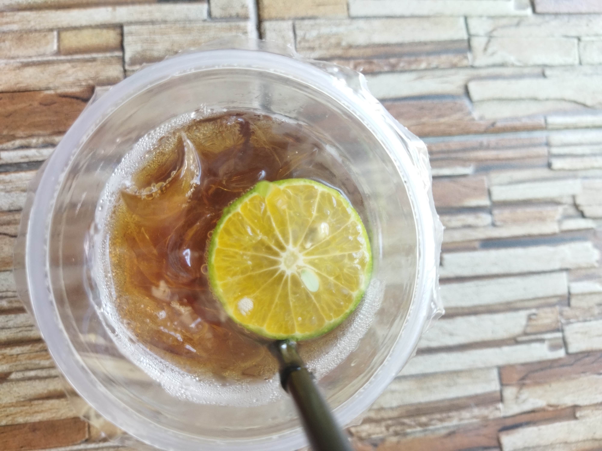A close-up, top-down shot shows a slice of lime floating on ice in a clear plastic cup filled with a dark beverage. The cup is placed on a surface with a textured, brick-like pattern, featuring shades of brown, beige, and gray. A black straw is visible, partially submerged in the drink and positioned beneath the lime slice. The background consists of this textured surface, extending to the right of the cup. The image does not depict any people or specific landmarks in Suci, Indonesia.