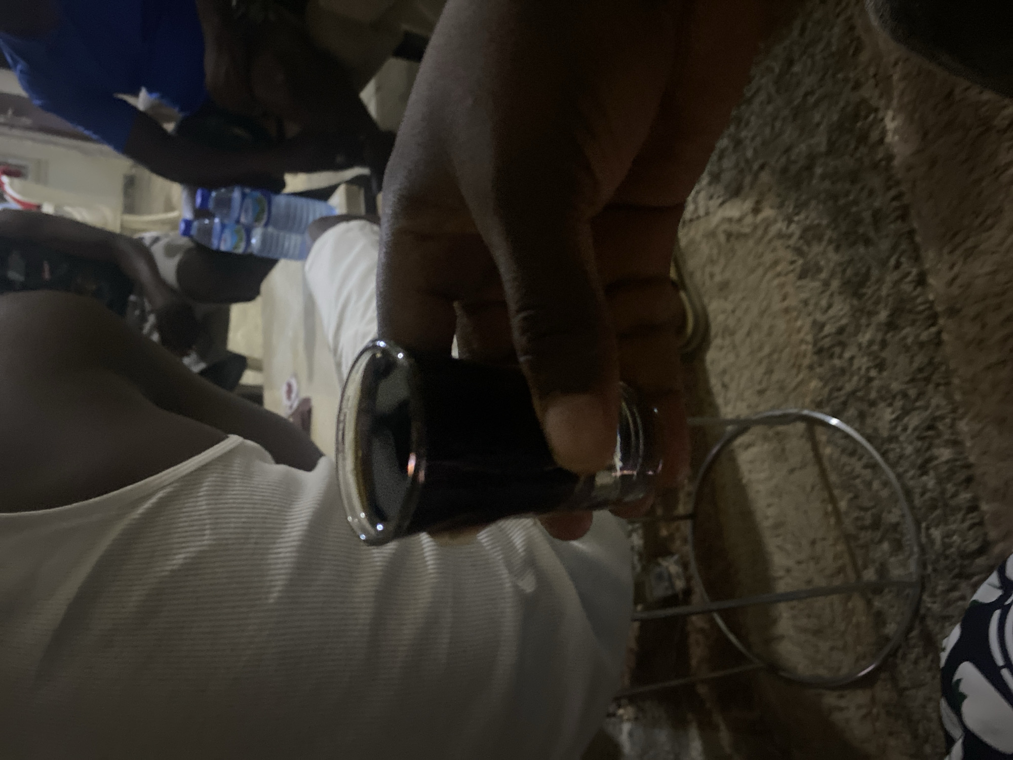 A dark-skinned hand holds a shot glass filled with a dark liquid. A person wearing a white shirt sits at a table, and other people are visible in the background. Plastic water bottles are also on the table. The location appears to be indoors, possibly in a social setting, given the arrangement of people and objects. The specific location within Barden Yana, Nigeria, cannot be determined from the image.