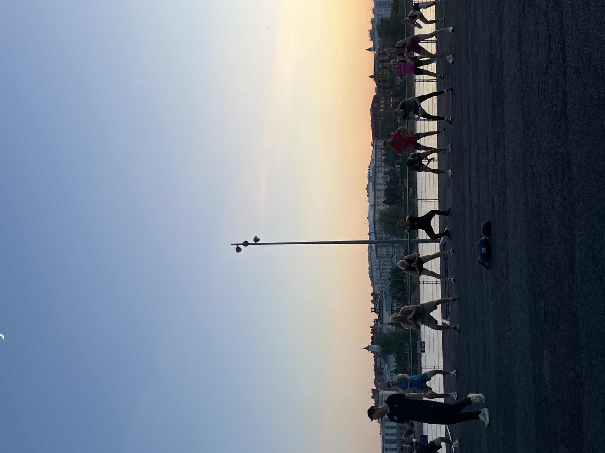The image shows a group of people doing a workout on a paved surface along the Kazanka River in Kazan, Russia. They are stretching and posing. The setting sun creates a gradient of color in the sky, with blue merging into orange near the horizon. Buildings, including the Kazan Kremlin, are visible in the background, across the river. A tall lamppost stands near the workout group.