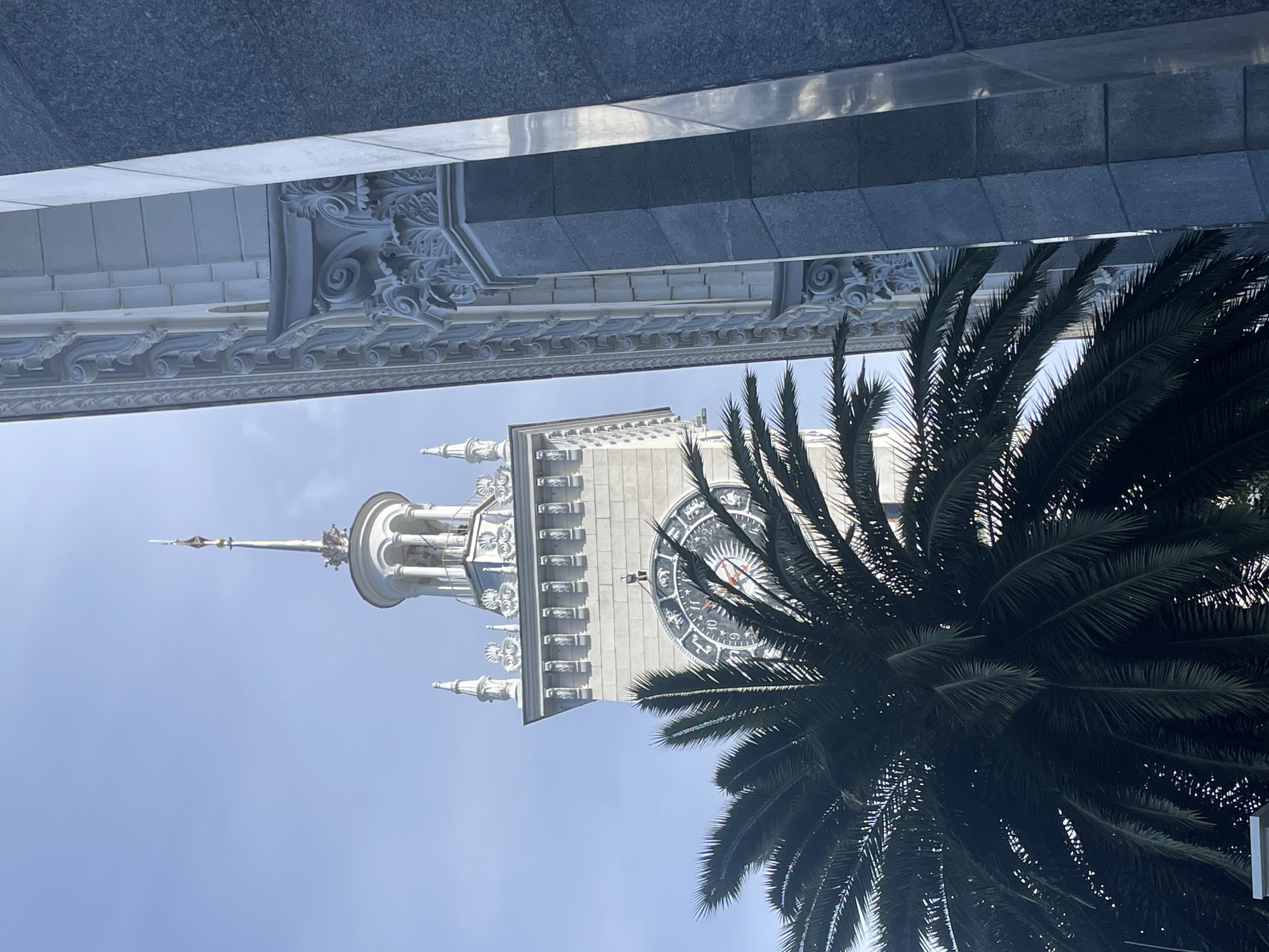 The image is an upward-angled shot featuring a tall, ornate clock tower partially obscured by a palm tree. The tower is light-colored, possibly white or light gray, with intricate architectural details, including multiple tiers, spires, and decorative elements. A clock face is visible on the tower, adorned with zodiac signs and other symbols. The palm tree, with its dense, dark green fronds, occupies the right side of the frame, partially covering the tower. The background consists of a clear blue sky. At the top of the image, a portion of a building with similar architectural detailing is visible, adding context to the urban setting. This is the Sochi Sea Port in Sochi, Russia.