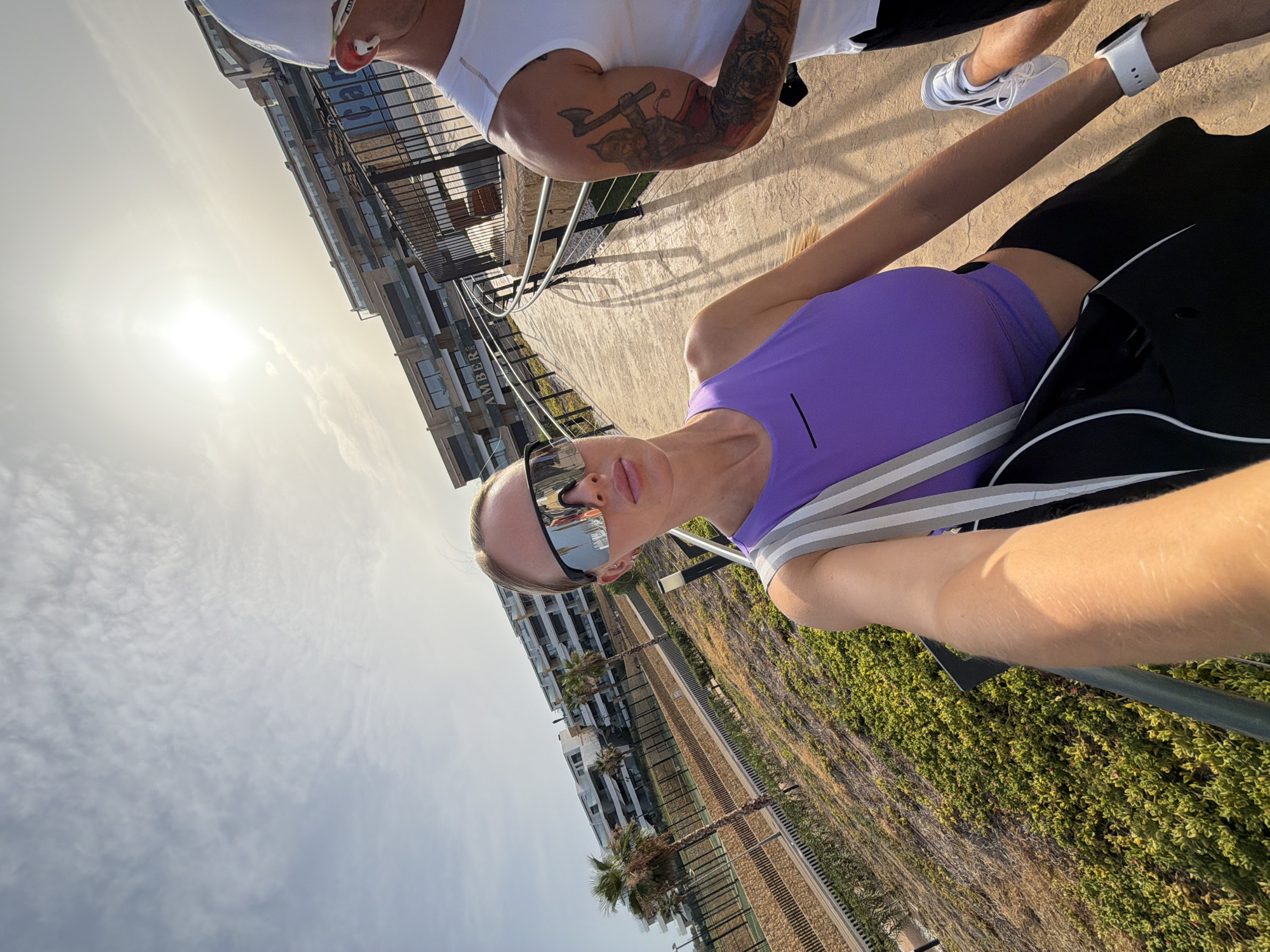 In Finestrat, Spain, under a bright, partly cloudy sky, a woman with blonde hair pulled back, wearing large reflective sunglasses, a purple tank top, and black athletic pants, holds a black bag with a gray and white strap. She is taking a selfie on a paved walkway lined with lush green vegetation and palm trees. To her right, a man with a white cap, white tank top, and visible arm tattoos is walking. In the background, a modern, multi-story building complex with balconies and windows is visible. The scene suggests an outdoor recreational activity, possibly exercise or a walk, in a contemporary urban or resort environment.