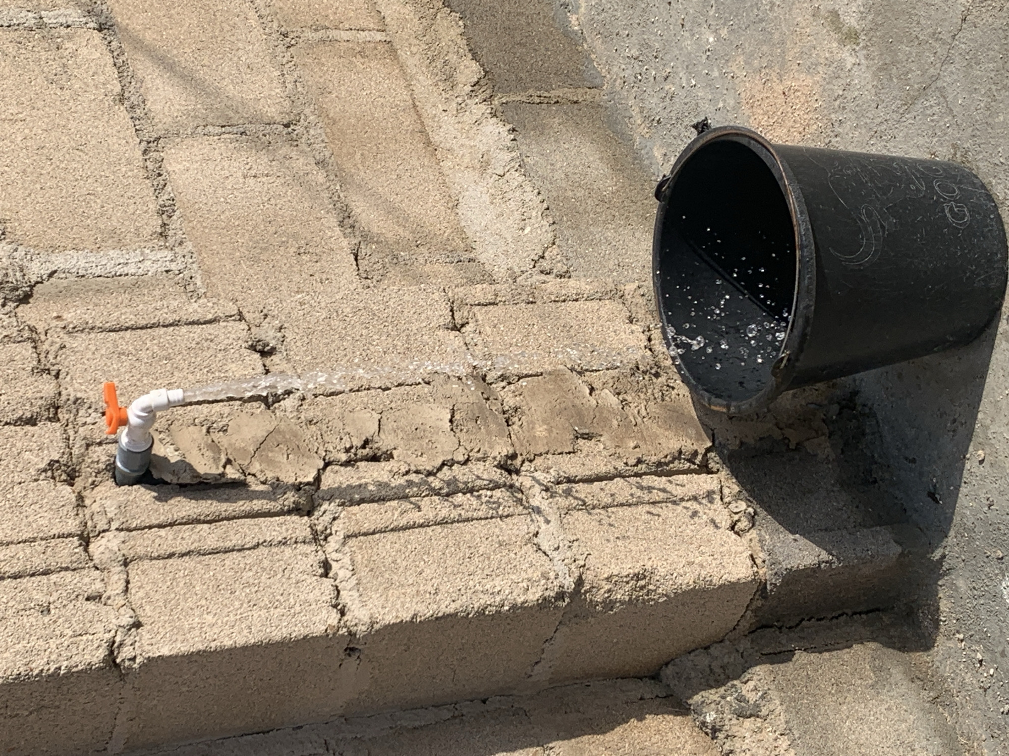An outdoor scene depicts a partially filled black bucket positioned on a concrete surface beneath a water tap affixed to a rough, light-brown cinder block wall. Water is flowing from the tap, creating a visible stream that falls into the bucket and splashes. The tap is white with an orange handle. The wall shows signs of age and weathering, with visible mortar lines and some cracking. Sunlight casts shadows on the wall and ground.