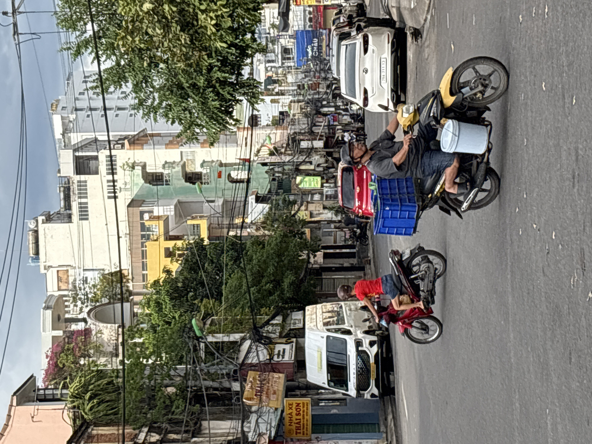 The image depicts a street scene in Nha Trang, Vietnam. In the foreground, two individuals are riding motorcycles. The first motorcycle, a red one, is ridden by a person wearing a red shirt and blue shorts. The second motorcycle, yellow and black, is ridden by a man wearing a dark grey short-sleeved shirt, blue denim shorts, and a helmet. He is transporting a blue plastic crate and a light blue bucket attached to the side of his motorcycle. In the background, a white van is parked on the left side of the street. Several cars, including a white sedan with the license plate "79A 711 13", are also visible. The street is lined with multi-story buildings, typical of urban architecture in Vietnam, with some adorned with plants and balconies. Overhead, a dense network of electrical wires crisscrosses the sky. The sky is overcast, suggesting potentially dull weather. The overall impression is one of a bustling urban environment with everyday traffic and street activity.