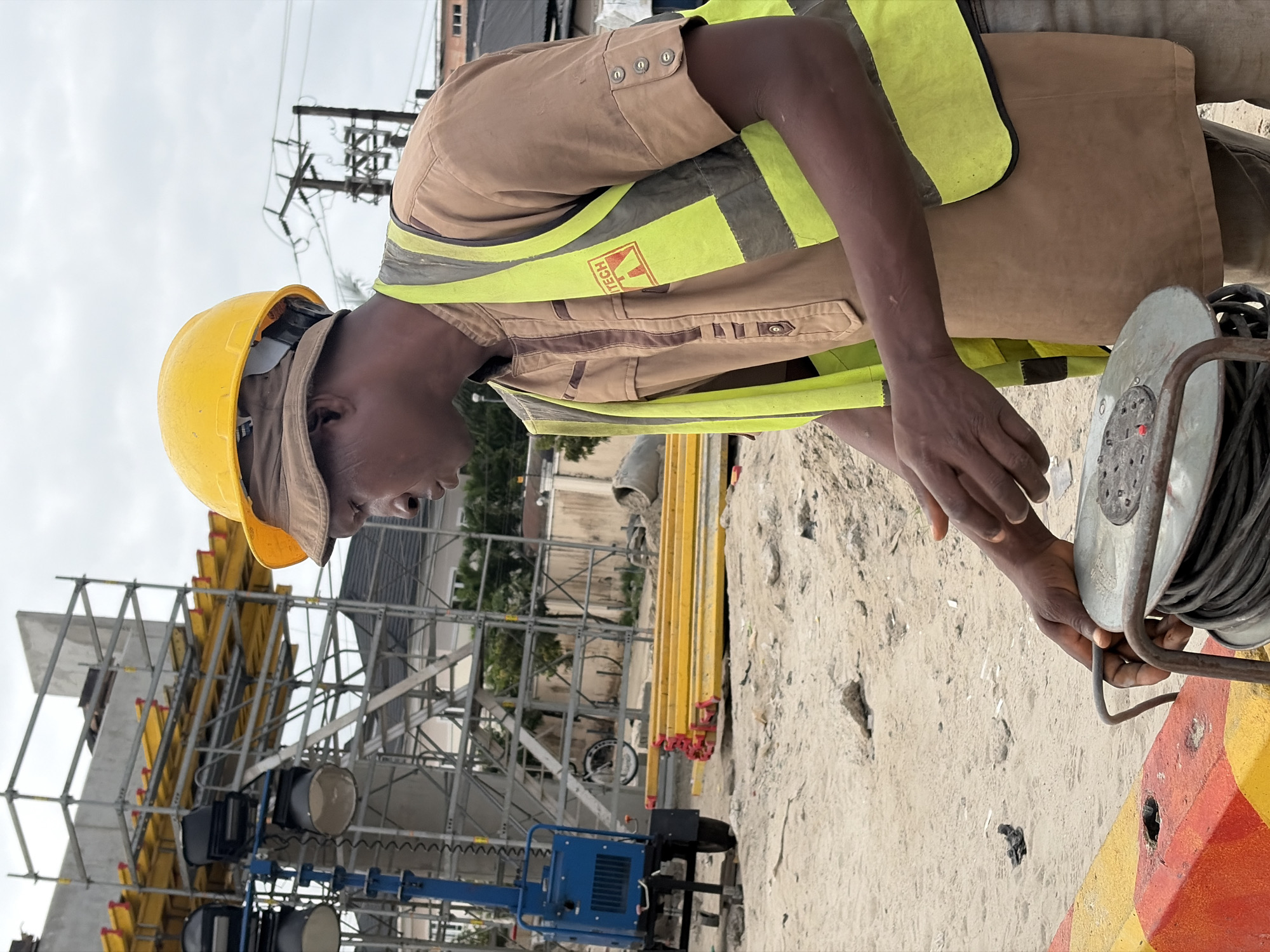 A man wearing a yellow hard hat, a brown garment, and a high-visibility vest is working at a construction site in Alaguntan, Nigeria. He is seen holding a coiled electrical cable. In the background, scaffolding, construction materials, and electrical poles are visible. The ground is sandy and appears to be part of the construction area.