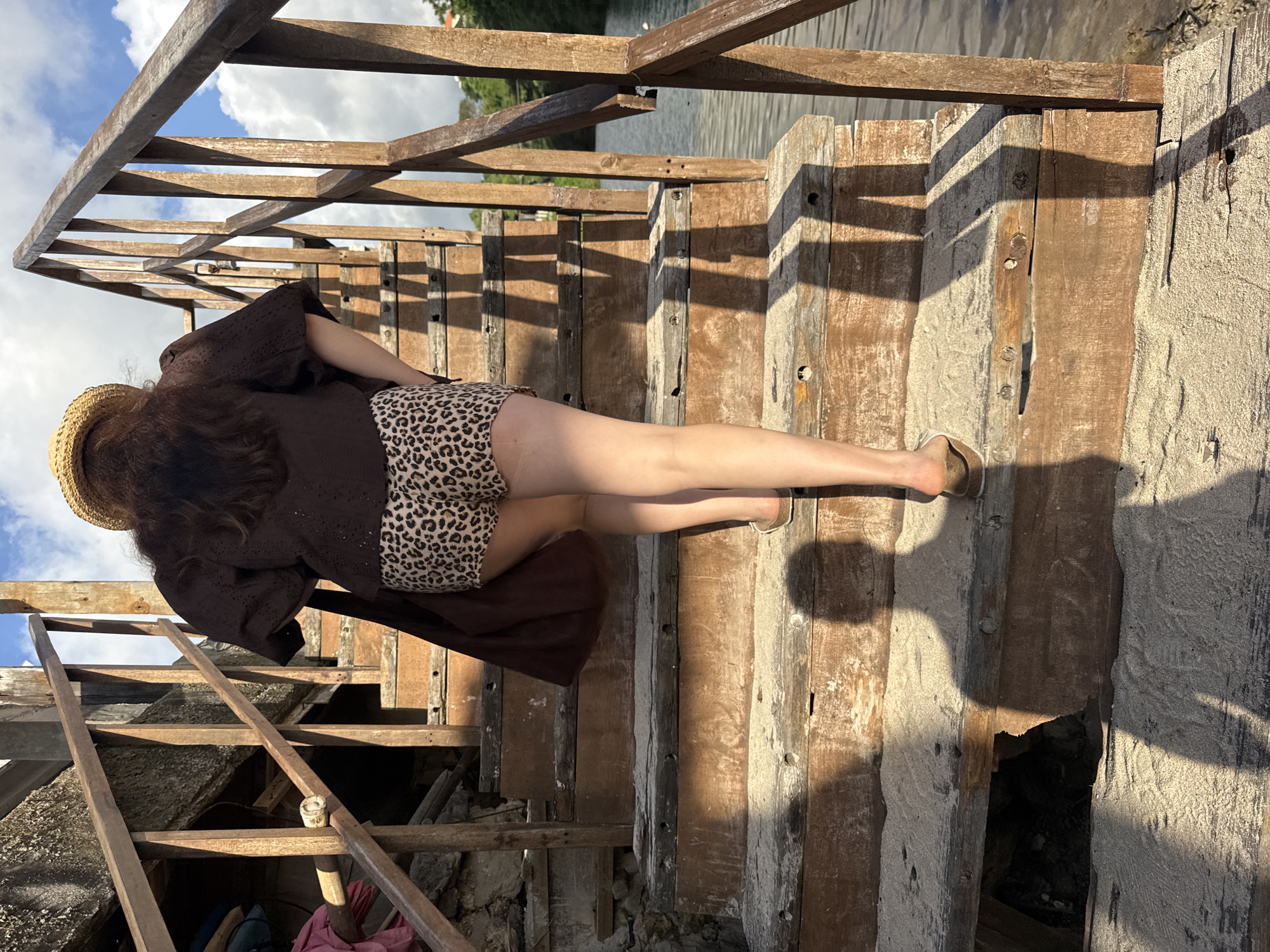 A woman stands on weathered wooden stairs near a body of water, likely a lake or ocean, in Kuta Selatan, Indonesia. She wears a straw hat, a brown, short-sleeved top, and leopard-print shorts. Her feet are clad in flat sandals. The wooden structure includes a railing, and shadows cast by the sun are visible on the steps and surrounding sand or sediment. Vegetation is visible in the distance.