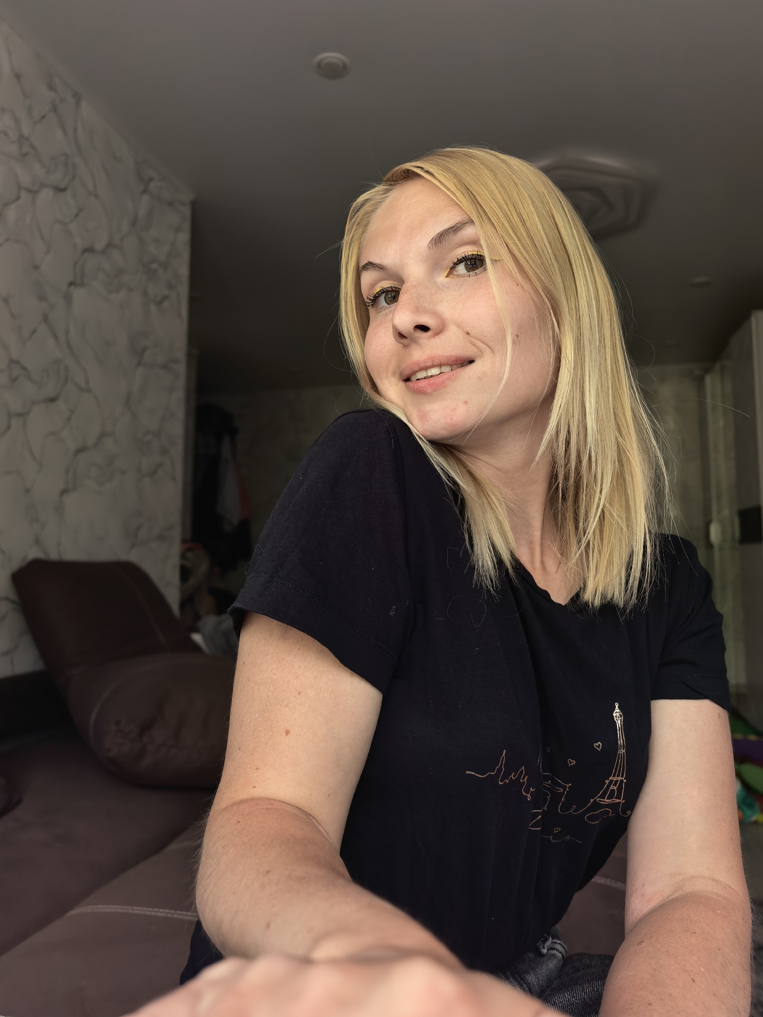 An indoor, medium-shot, eye-level, selfie-style photograph shows a young adult Caucasian female with blonde hair and yellow eyeliner. She is wearing a black t-shirt with a subtle, metallic graphic of the Eiffel Tower. She is looking towards the camera with a slight smile. The background includes a patterned wall, a brown armchair or sofa, and a hallway with what appears to be clothing hanging on a rack. The lighting suggests an indoor environment, possibly a home. No specific location in Sterlitamak, Russia can be determined from the visual information.