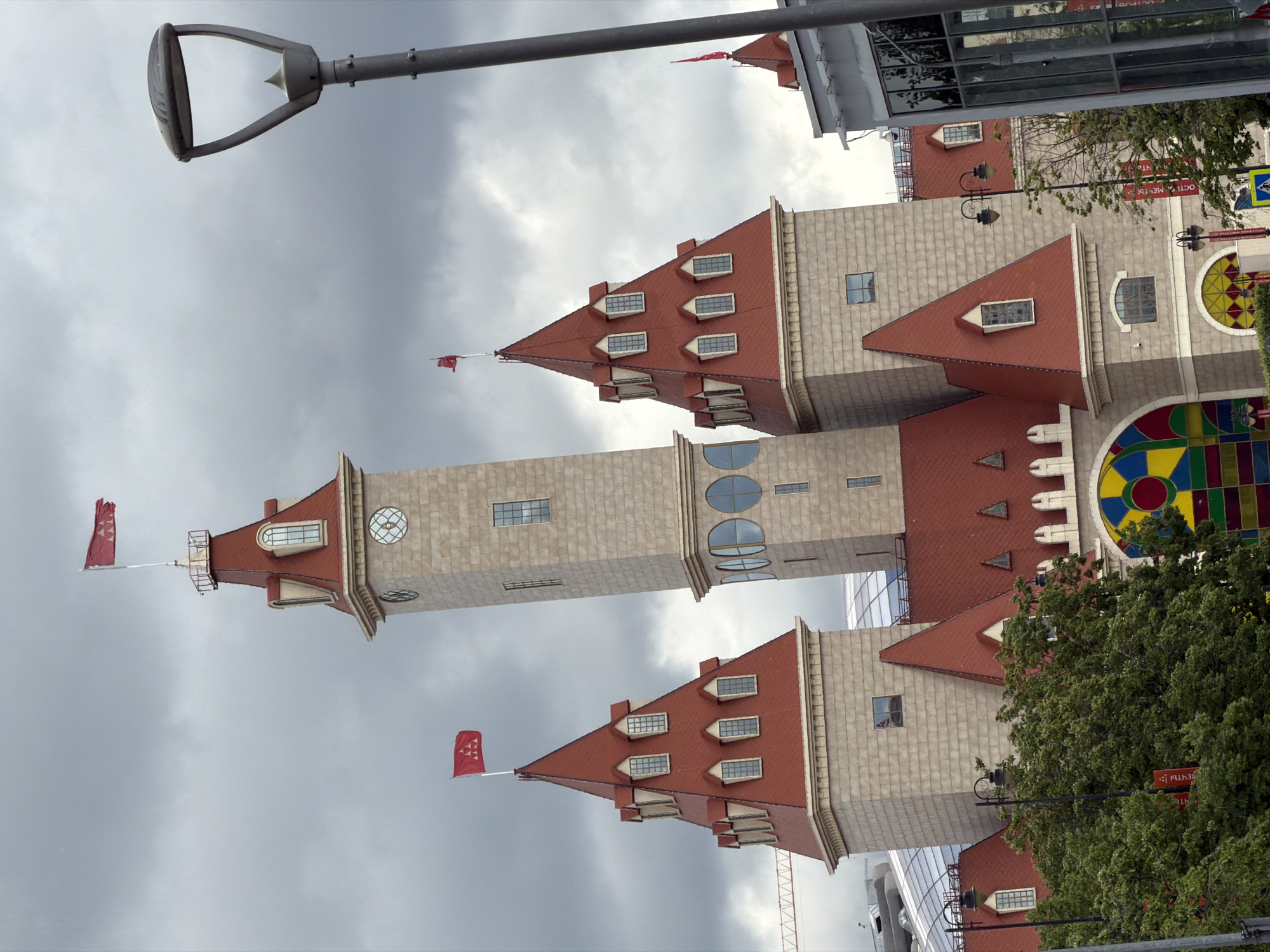 The image shows a building with multiple towers and red roofs against a cloudy sky. Red flags with white patterns are visible atop some of the towers. The building features elements of modern architecture with geometric designs. A streetlight is in the foreground. Based on the architecture and the presence of flags that match historical Moscow flags, the location is likely in the city of Moscow, Russia.