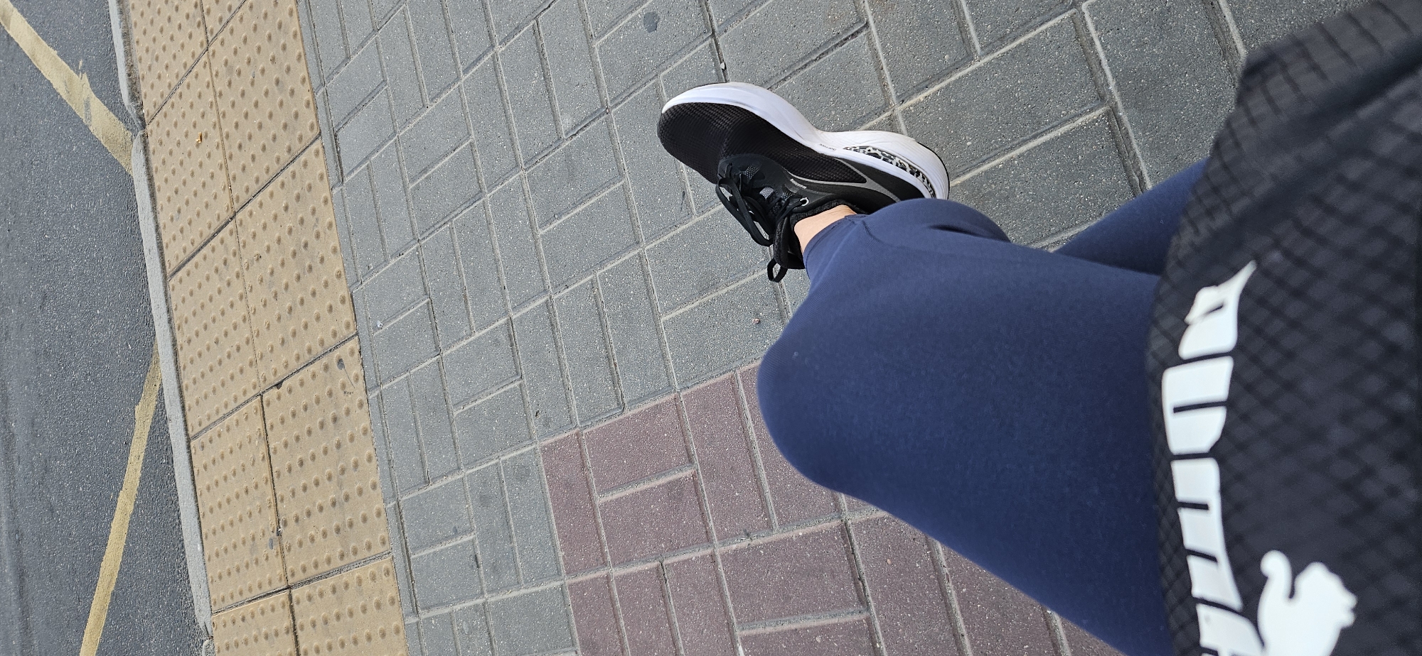 The image shows a person's legs, wearing dark blue leggings and a black sneaker with a white sole, sitting on a pavement. The pavement is made of rectangular tiles of different colors and textures. The background shows a road with a yellow line and tactile paving tiles. The scene is outdoors, on a sunny day. It is not possible to determine the specific location in Vitsebsk, Belarus based solely on the visual information provided.