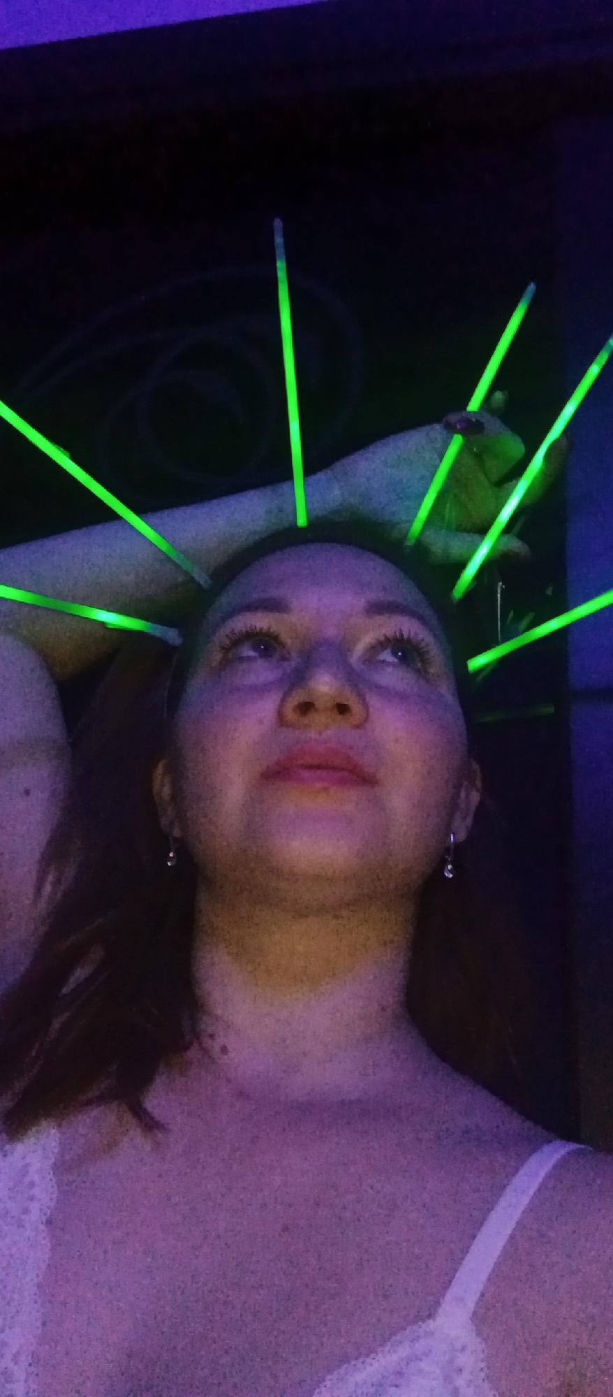 A close-up, low-angle shot shows a person illuminated by neon lights, facing upward. The individual, likely female, has fair skin, reddish-brown hair, and is wearing small hoop earrings. Multiple green glow sticks are emanating from the top of their head, resembling a crown. A hand, presumably belonging to another person, is visible in the upper right of the frame, also adorned with a green glow stick. The setting appears to be a dimly lit interior, with violet and black hues dominating the background. The person is wearing a white garment with lace on the shoulder strap.