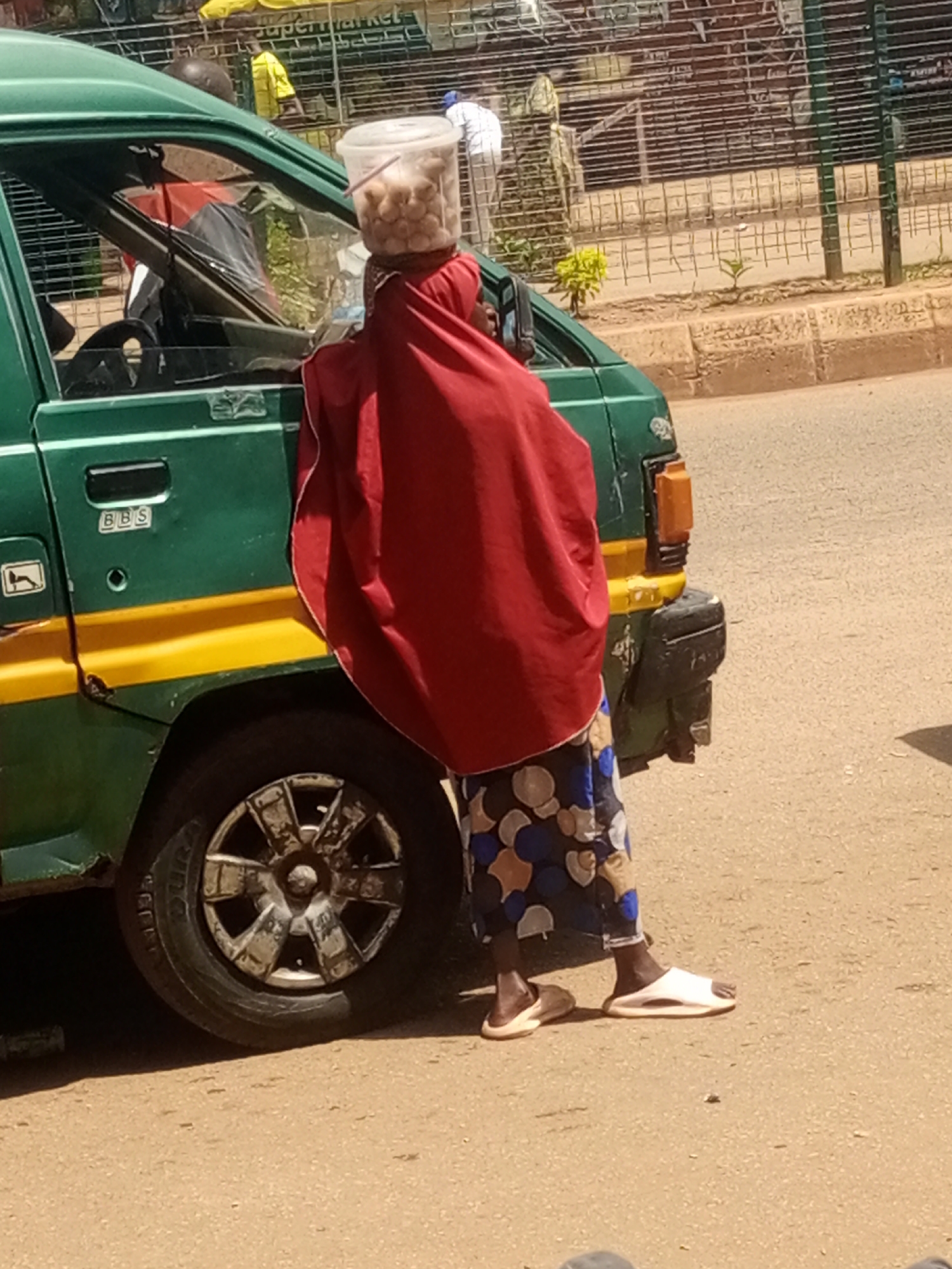A person, covered in a red cloth, stands next to the open passenger door of a green and yellow van. The person is holding a clear plastic bucket filled with what appear to be potatoes. A straw is sticking out of the bucket. They are wearing a patterned skirt and white slide sandals. The van has a "BBS" logo on its side. The scene appears to be on a dirt or gravel road next to a sidewalk with a metal fence in the background. Beyond the fence, a yellow-clad figure and other people are visible, along with a sign that reads "SUPERMARKET." The time of day appears to be daylight, possibly mid-morning or afternoon, with the sun casting shadows. The overall setting suggests a market or roadside vending area.