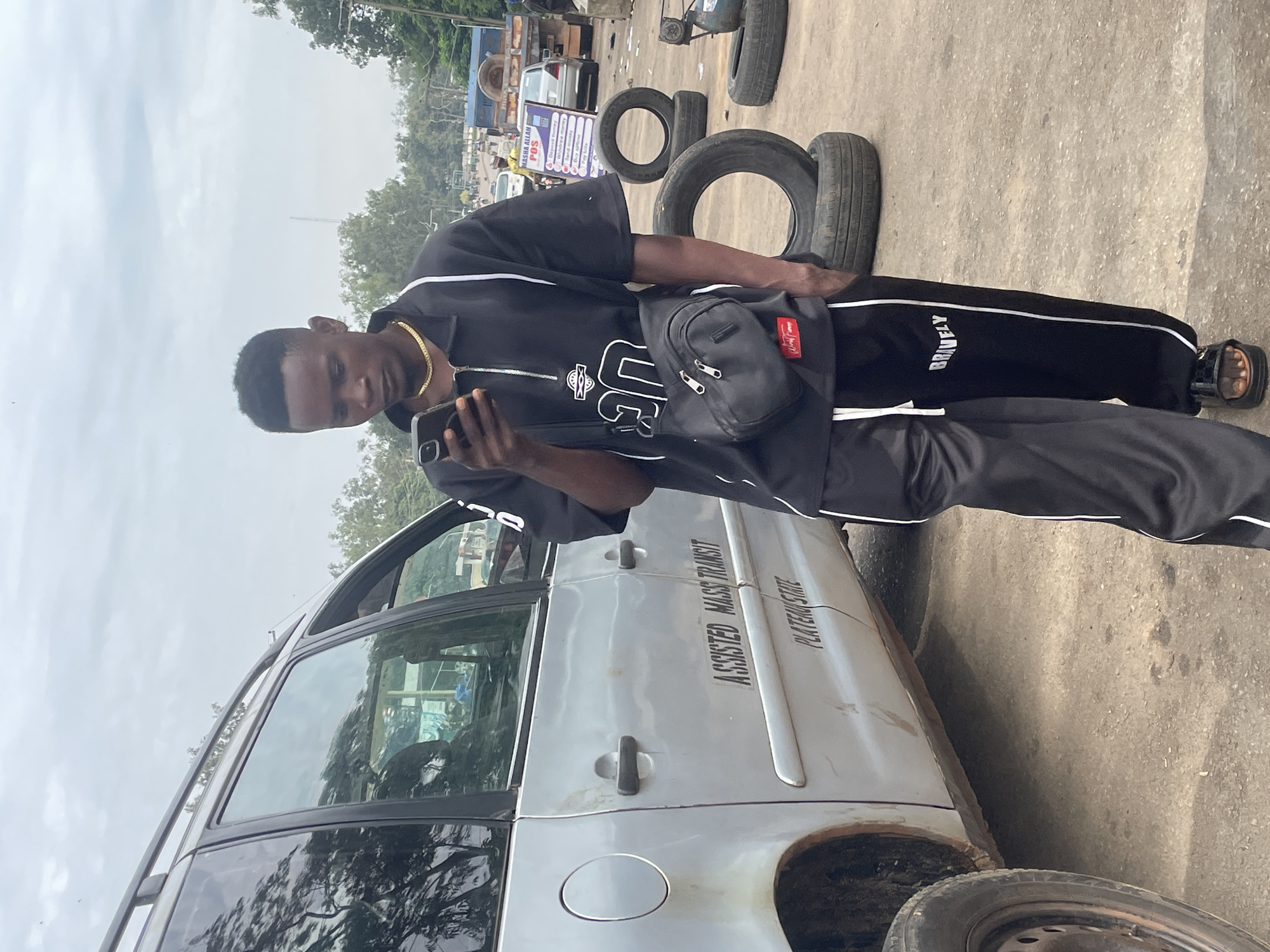 A young Black male is standing next to a silver minivan in an outdoor urban setting. He is wearing a black zippered shirt with white accents and black pants with white stripes. He is holding a smartphone and looking down at it. The minivan has the words "ASSISTED MASS TRANSIT" and "PLATEAU STATE" written on its side. Several old tires are stacked behind him. In the background, there are other vehicles, including trucks, and a sign that reads "MASHA ALLAH POS." The ground is paved and appears to be a road or parking area. The sky is overcast. This scene is consistent with a location in Jos, Nigeria, a city known for its mass transit services and busy urban environments.