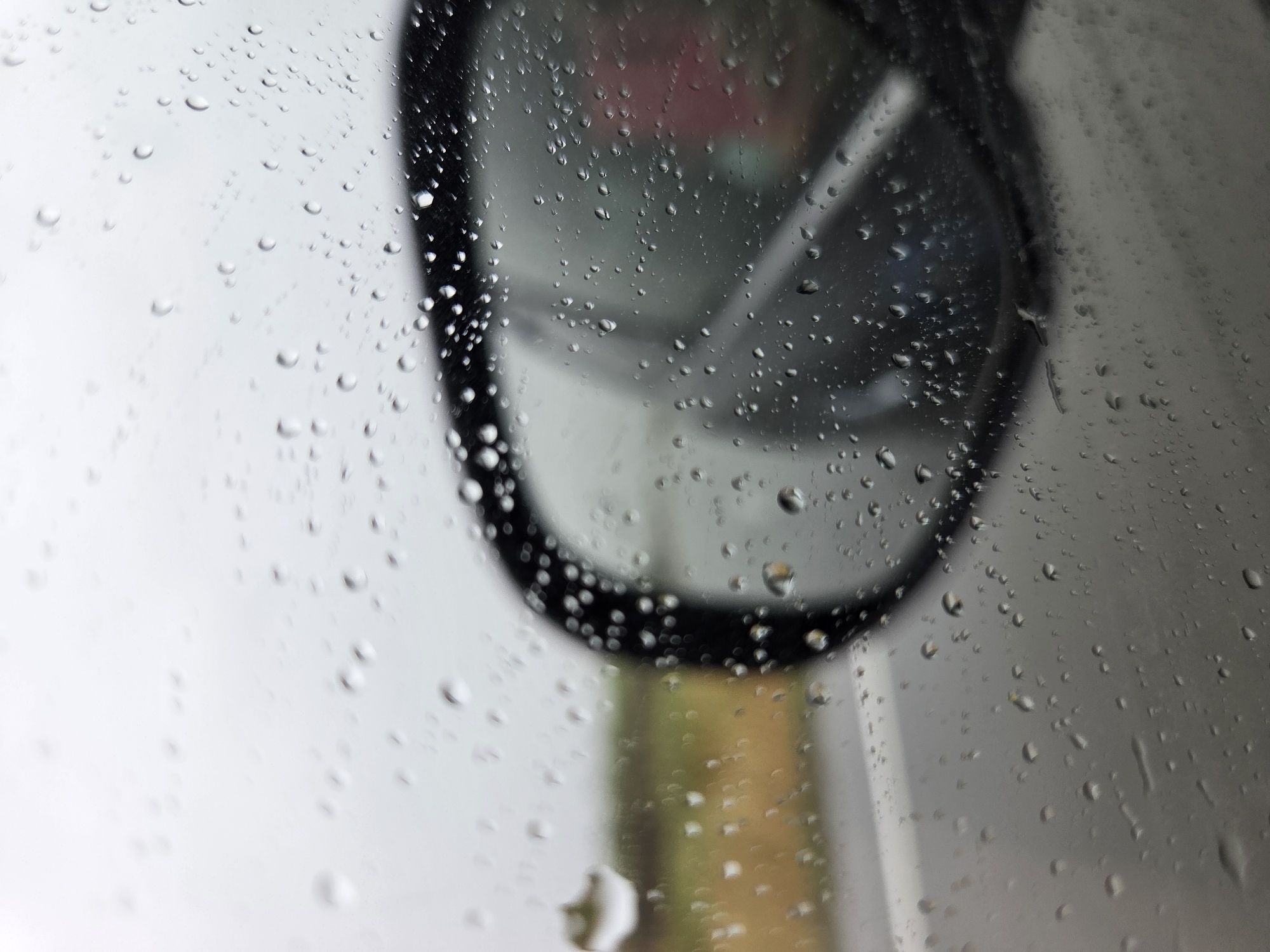 The image captures a close-up, out-of-focus view of numerous raindrops clinging to a glass surface, likely a car window. The droplets vary in size and shape, creating a textured foreground that blurs the elements behind them.
In the mid-ground, slightly out of focus, is the dark, oval-shaped frame of what appears to be a vehicle's side mirror or rearview mirror. The surface of the mirror itself reflects soft, indistinct colors, including hints of pink, green, and a muted yellow or brown, suggesting some distant objects or landscape are being mirrored. The light background around the mirror suggests an overcast sky or diffused daylight.
The scene depicts a moment during or immediately after rainfall, viewed from inside a vehicle. There is no specific activity taking place, but the image conveys the atmosphere of a rainy day from an interior perspective. No people are visible.
Notable details include the crispness of the individual water droplets contrasted with the soft, blurry background, emphasizing the glass as the primary focal point. The overall lighting is subtle and diffused. The provided location context is Бабстово, Russia, though no specific visual cues within the image confirm this location. There is no visible text.