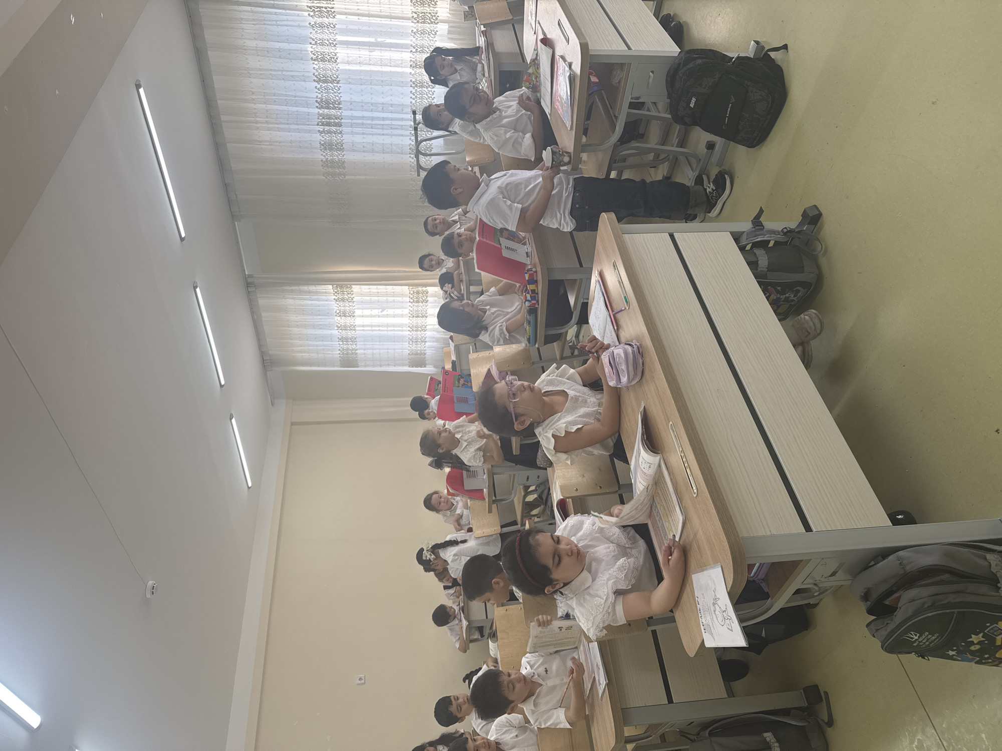 The image depicts a lively classroom scene in Tashkent, Uzbekistan, filled with young primary school students during what appears to be daytime. The classroom is arranged with multiple rows of individual wooden-topped desks and chairs with light grey metal frames.
Numerous children, predominantly wearing white shirts – some with decorative ruffles or lace – and dark bottoms, are seated at their desks. They appear to be engaged in a learning activity, with many students looking at open books, notebooks, or papers on their desks, suggesting a reading or writing lesson. Their expressions are generally focused and attentive.
The room is well-lit by several long, white fluorescent strip lights on the ceiling and abundant natural light entering through large windows, which are covered with sheer, patterned white curtains. The walls are a light cream or white, and the floor is a pale yellow-green.
Various objects are visible, including backpacks placed under desks or on the floor. One backpack prominently displays the Ukrainian trident symbol and the Cyrillic word "УКРАЇНА" (Ukraine). Another backpack has "SABER SPORTS" or "SAGAR SPORTS" written on it. On one desk, a paper shows an outline drawing of a frog with the Cyrillic word "ЖАБА" (frog). Other visible text on books also appears to be in Cyrillic script. A wall-mounted white device, possibly a smoke detector or sensor, and an electrical outlet are also present on the walls. The overall setting conveys a typical, active school environment.