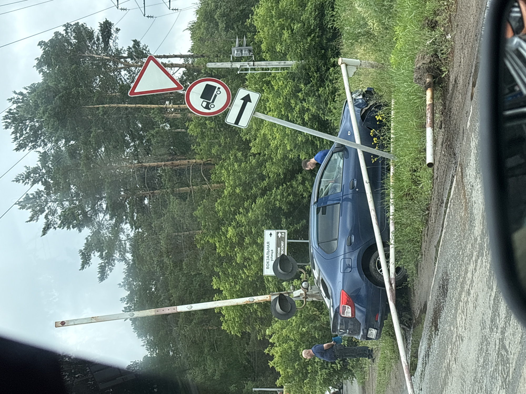 The image depicts a traffic incident at a railway crossing in Chaikovsky, Russia. A blue sedan has collided with the railway crossing barrier, causing damage to the vehicle's front end and bending the barrier. The barrier arm lies on the ground, partially submerged in mud and water. Two men are present near the scene. One man, wearing a blue shirt and dark pants, stands by the side of the road with a blue object in his hand, seemingly observing the situation. Another man, wearing a blue t-shirt, is seated inside the damaged car. Several traffic signs are visible, including a yield sign, a sign prohibiting trucks, and a directional arrow indicating to turn right. The environment is characterized by lush green trees and foliage, with overcast skies overhead. Power lines are also present in the background. The road surface appears wet and muddy, suggesting recent rainfall.