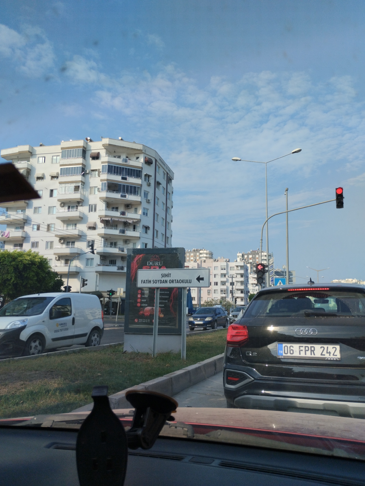The image shows a street scene in Mezitli, Turkey, with a multi-story white apartment building in the background. A black Audi Q2 with license plate "06 FPR 242" is in the foreground, stopped at a traffic light. A white van is visible to the left of the Audi. A street sign indicates directions to "ŞEHİT FATİH SOYDAN ORTAOKULU". The sky is blue with scattered clouds. The scene appears to be a typical urban environment during daylight hours.