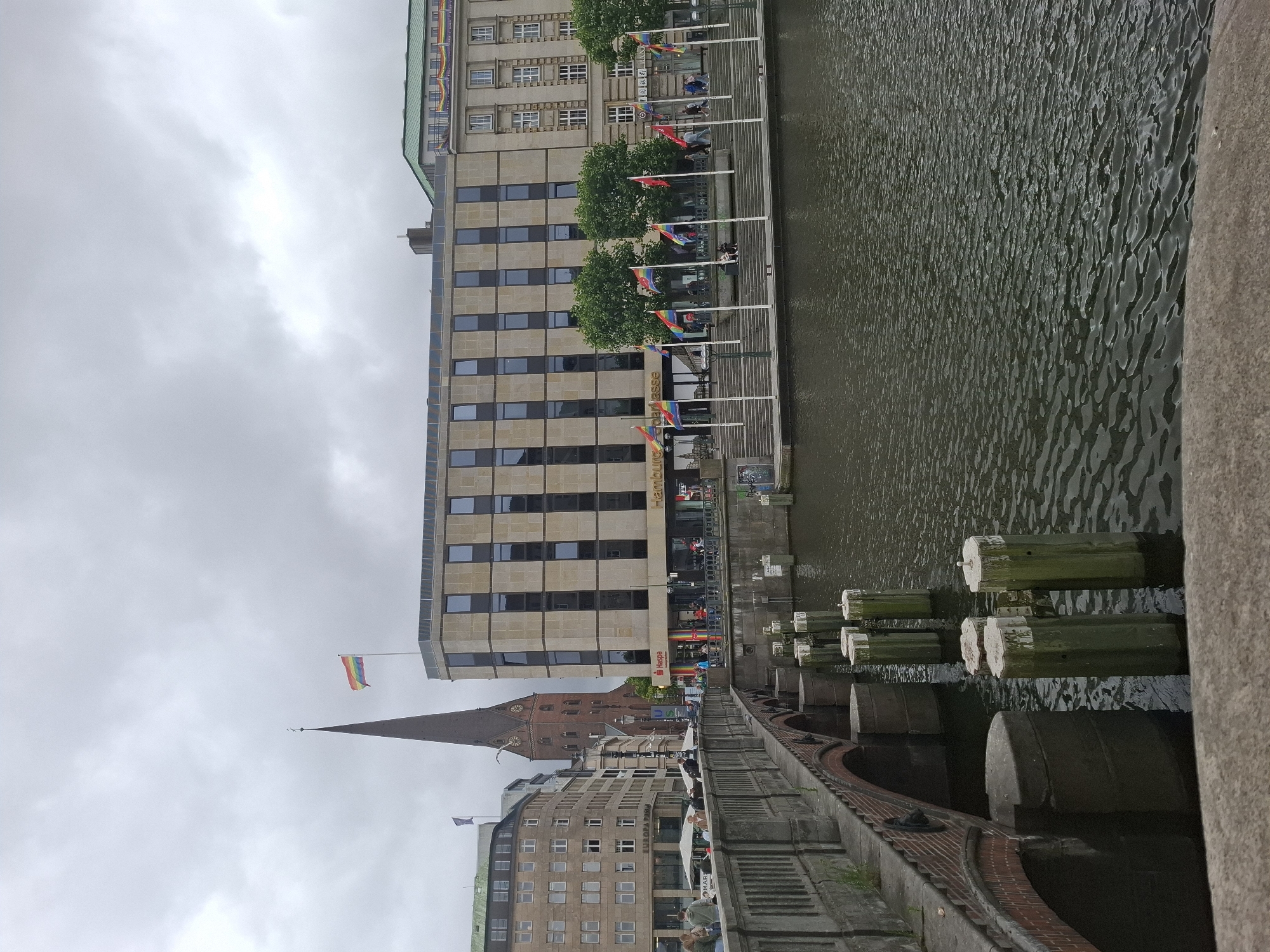 The image captures a scene in Hamburg, Germany, specifically along a canal. In the foreground, a stone bridge with red brick archways spans the water, with several concrete posts emerging from the dark, rippling water. The bridge is lined with a low concrete barrier.
Beyond the bridge, a modern building with a light-colored stone facade and rows of dark windows stands prominently. A rainbow flag is visible on top of this building and also displayed on poles along the waterfront in front of it. To the left of this building, a tall, dark-colored church spire with a clock face rises against a cloudy sky. Further to the left, a multi-story building with a light facade and many windows is visible.
Several people are walking and standing along the waterfront in front of the modern building. The environment is overcast, suggesting a cloudy day. The scene appears to be a public area with a mix of historical and modern architecture.