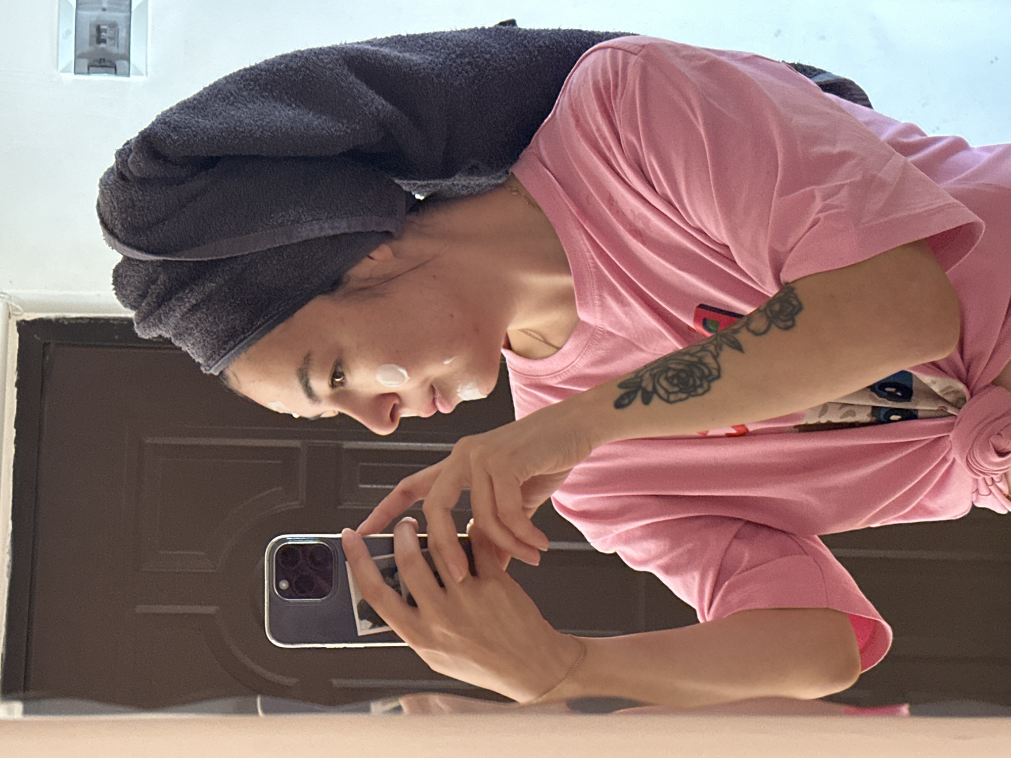 A young woman is taking a mirror selfie in what appears to be a bathroom. She has a grey towel wrapped around her head, and white cream applied to her face, likely for skincare. She is wearing a pink t-shirt tied at the waist and has a tattoo on her left arm. The background shows a brown door and a white wall with a light fixture. The image does not provide any specific visual cues to indicate the location is Gyumri, Armenia.