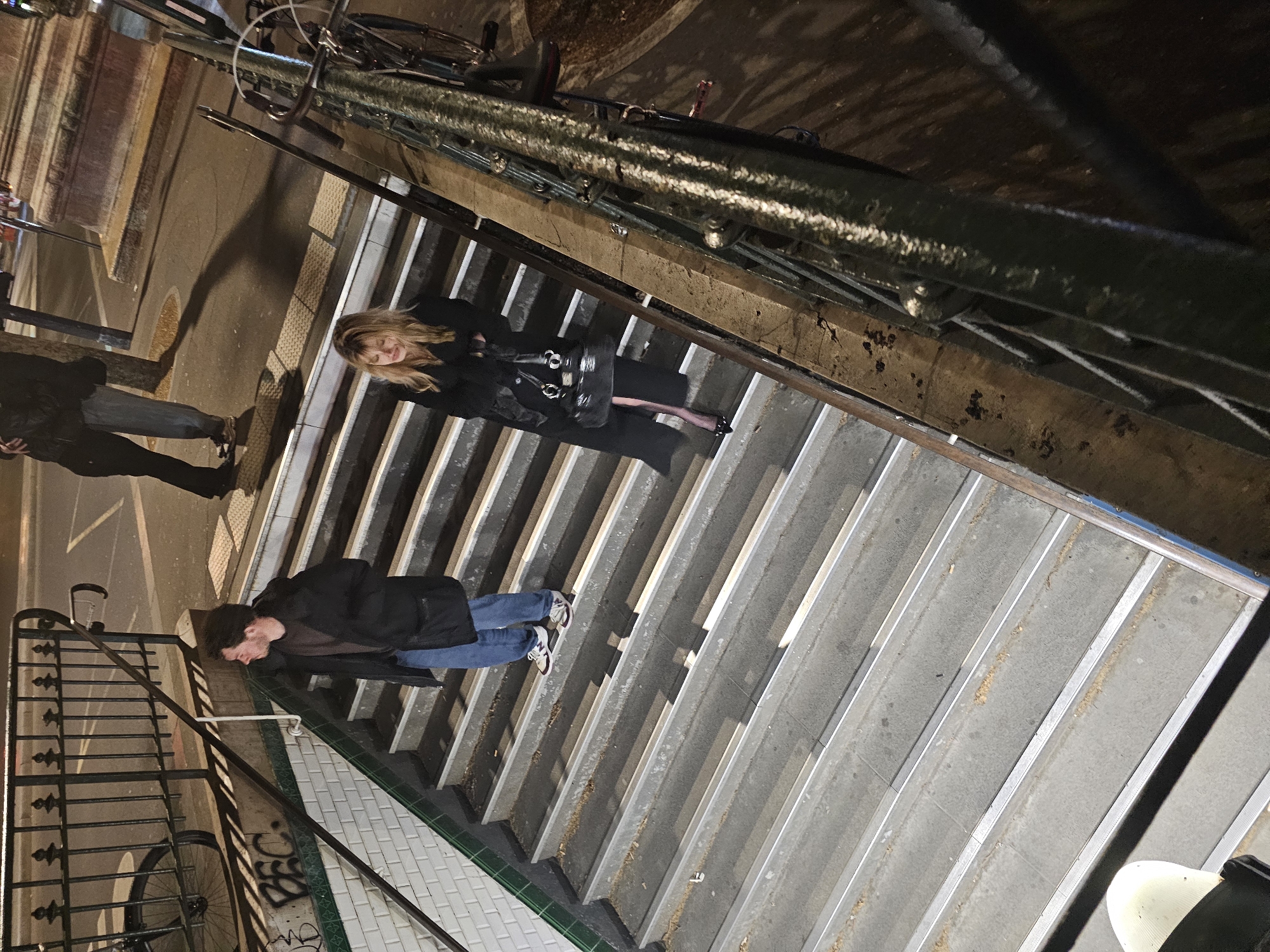 Here's a forensic description of the image:
**Location:** Appears to be a Parisian subway entrance, indicated by the white tiling with a green border, graffiti on the tile, and the design of the metal handrail.
**Subjects and People:**
* Two pedestrians are descending the stairs, a woman in a dark, long coat and boots, and a man wearing a dark jacket and jeans.
* A third person can be seen in the background, standing on the street near the subway entrance.
**Objects:**
* Metal railing and stairs of the subway entrance.
* A bicycle is parked above the subway stairs.
* A woman is carrying a dark colored handbag.
**Actions:**
* The man and woman are descending the stairs.
**Environment:**
* Lighting: Low, likely nighttime.
* The environment is urban with a street alongside the subway entrance.