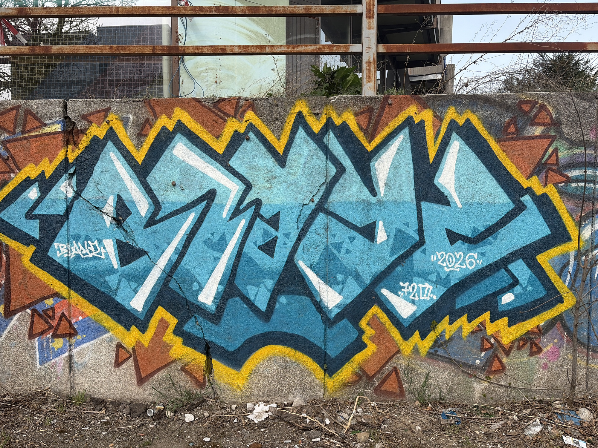 A concrete wall is covered in graffiti. The graffiti is largely blue with white accents and outlined in black. A brown and yellow jagged border surrounds the blue graffiti. There is additional brown graffiti in the form of triangles. Above the wall is metal infrastructure. The bottom of the wall is close to the ground, and the ground consists of dirt and trash. A small bare tree is to the right. The graffiti reads "RZA". The year "2026" is written in white above "7217". The location is likely in Sofia, Bulgaria.