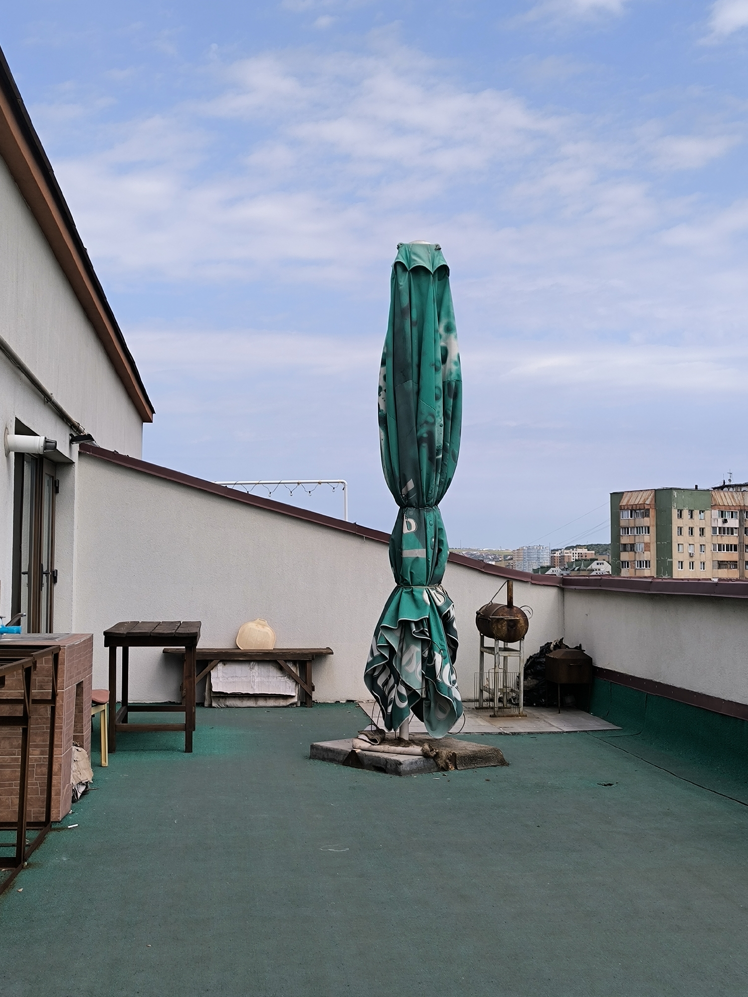 This is a photograph taken on a rooftop terrace in Chișinău, Moldova. The terrace has a green, textured surface. In the foreground, a large, folded green umbrella with a white pattern stands upright. To the left, there is a wooden table and a bench with a light-colored object on it. In the background, a white building with a brown-trimmed roof is visible on the left, and apartment buildings can be seen in the distance to the right. The sky is partly cloudy and blue. There are no people visible in the image.