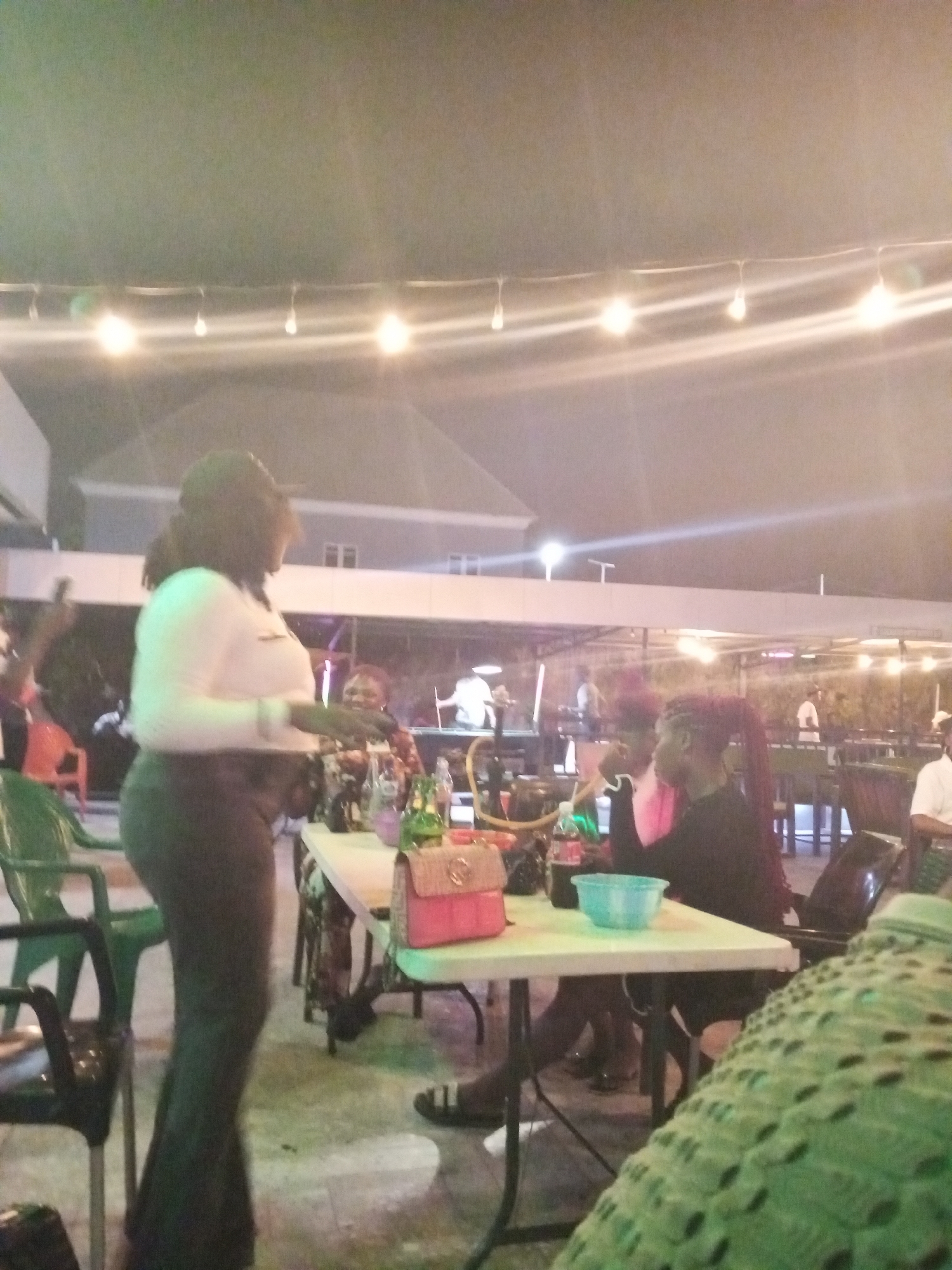The image shows an outdoor gathering at night, likely in a public or commercial space. In the foreground, a woman in a white top and dark pants stands to the left of a table. Two people are seated at the table. One woman has braids. The table holds various objects including bottles, a red purse, and a blue bowl. Additional tables and chairs are in the background, along with other individuals. String lights are strung overhead. The location is Adankolo, Nigeria.