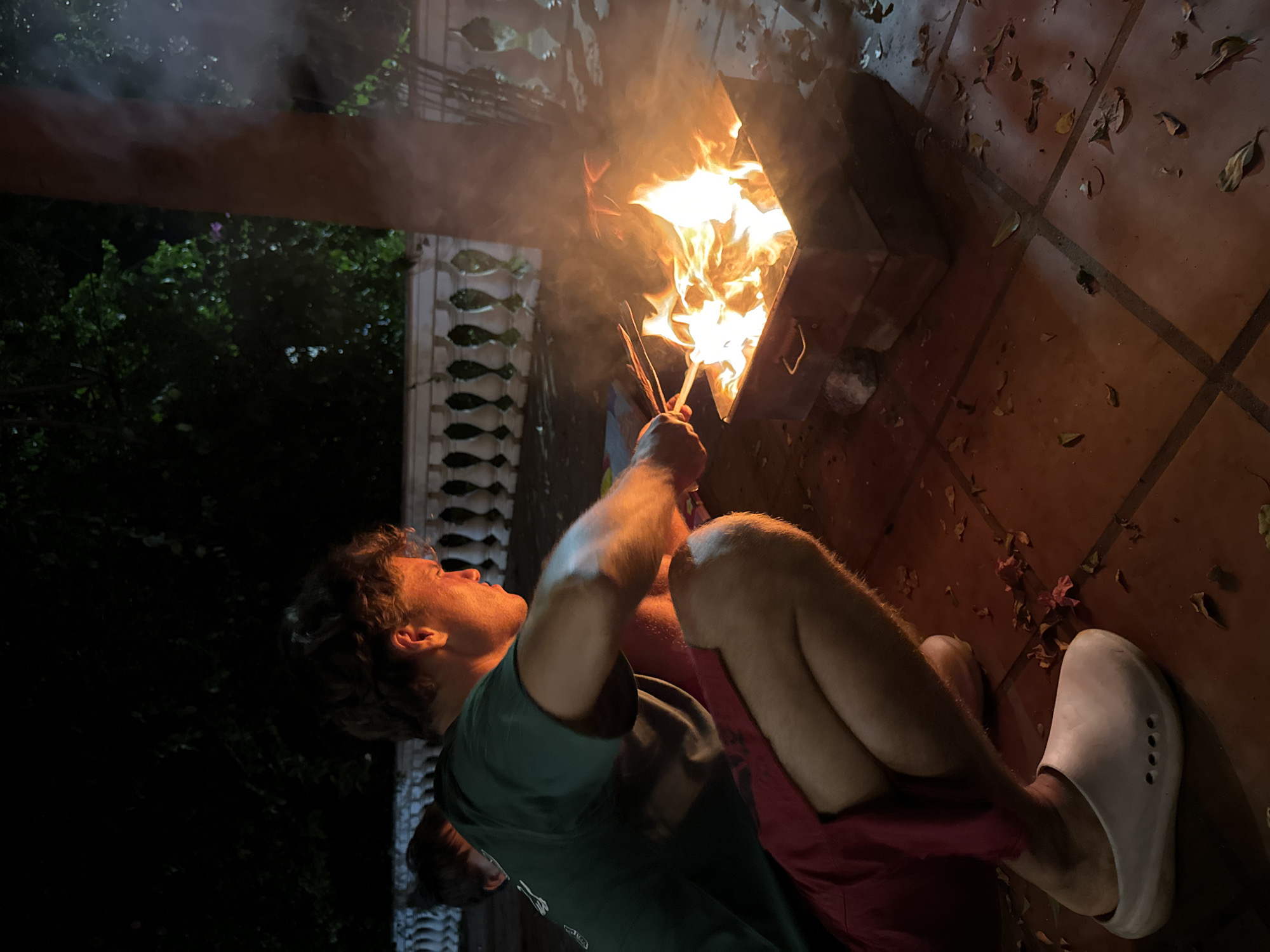 A young man is crouching on a tiled patio in front of a metal brazier filled with a bright, roaring fire. He is wearing a green t-shirt and red shorts, and white clogs. He is holding several sticks, which he appears to be using to tend the fire. Smoke rises from the fire and drifts to the right. In the background, a white ornate railing runs along the edge of the patio, and beyond that, dark foliage is visible against a night sky. Fallen leaves are scattered across the patio tiles. The scene suggests an outdoor gathering at night in Mũi Né, Vietnam, likely for a barbecue or bonfire. Another person's shoulder is visible to the left of the main subject.