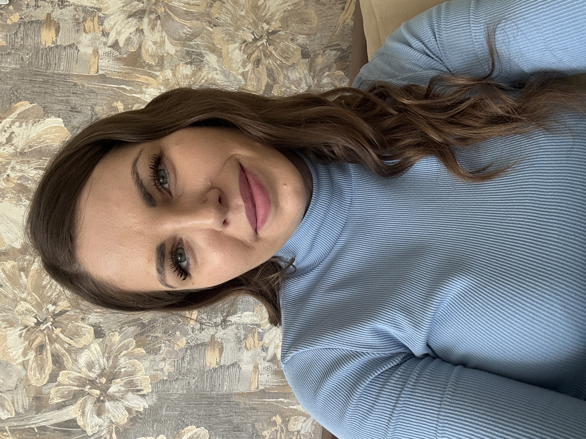 A woman is seen in a close-up shot wearing a light blue ribbed turtleneck sweater. She has long, brown hair with loose curls and green eyes. Her skin tone is fair, and she has pink lipstick on. The background features a wallpaper with a floral pattern in neutral tones. The image does not include any definitive markers to confirm the location in Раздолье, Russia.