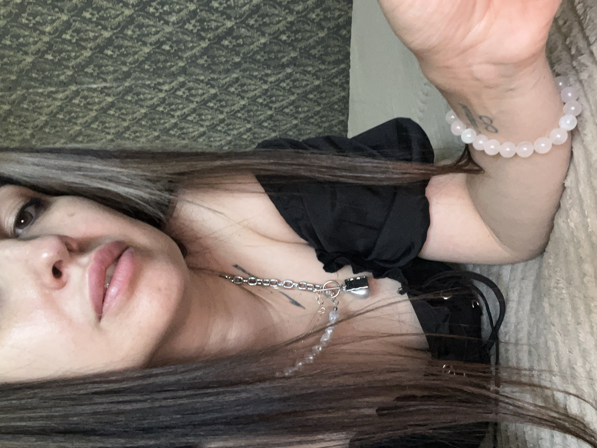 A woman is lying down, facing the camera, in an indoor setting. She is wearing a black garment, and has a silver chain with a pendant around her neck. A bracelet of light pink beads is on her right wrist. There is a tattoo on the underside of her wrist. Her hair is long and dark. The background includes a wall and a soft, textured surface. The city and specific location are unknown.
