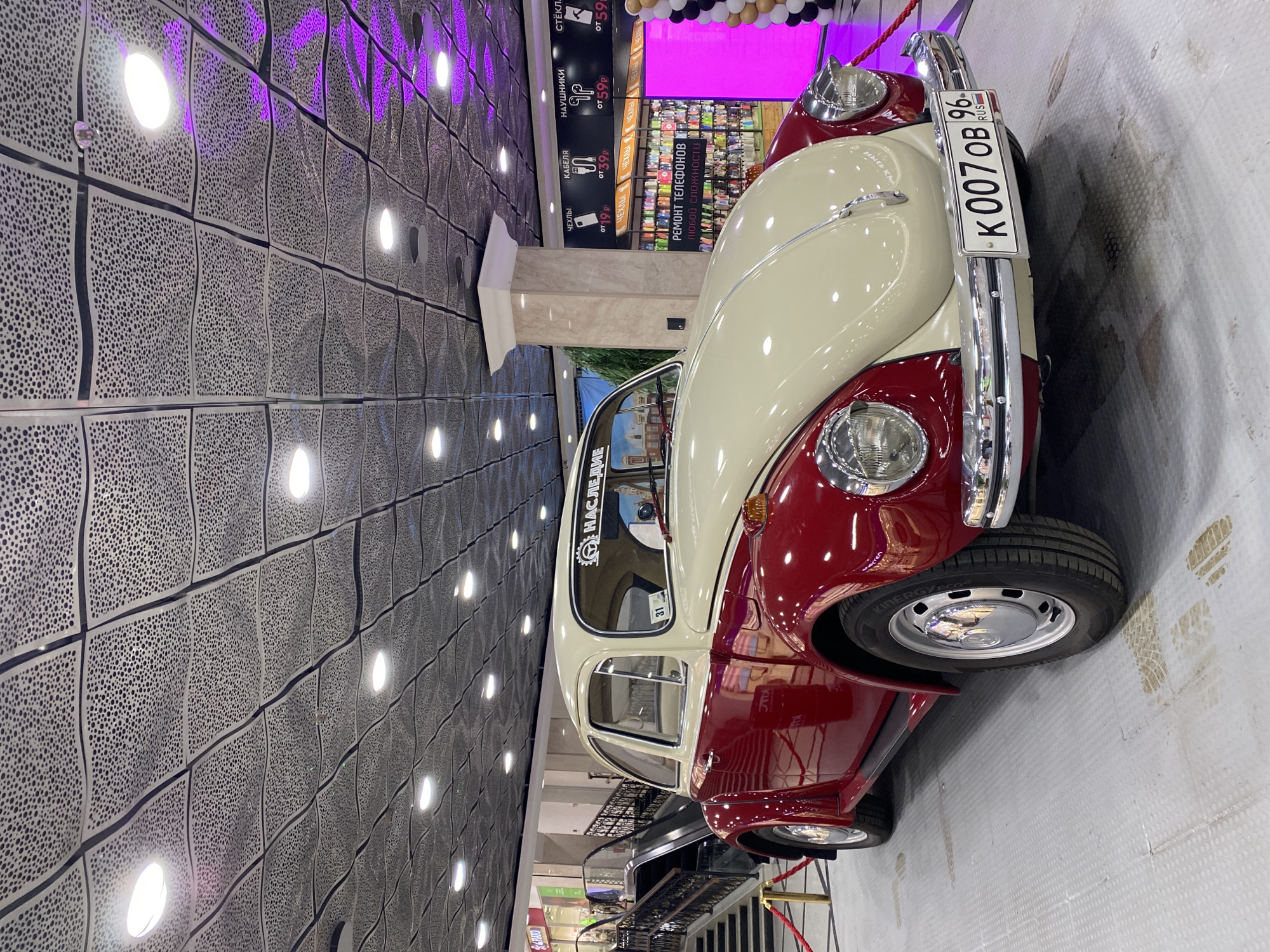 The image shows a red and white two-door Volkswagen Beetle parked indoors. The car has a license plate that reads "K 007 OB 96 RUS", indicating it is registered in Russia. Above the car, there is an advertisement for phone repair services and accessories such as cables, headphones, and screen protectors. The ceiling has a patterned design with integrated lighting. A staircase is visible in the background, along with a red rope barrier. The car is situated in a shopping mall located in the city of Yekaterinburg, Russia.