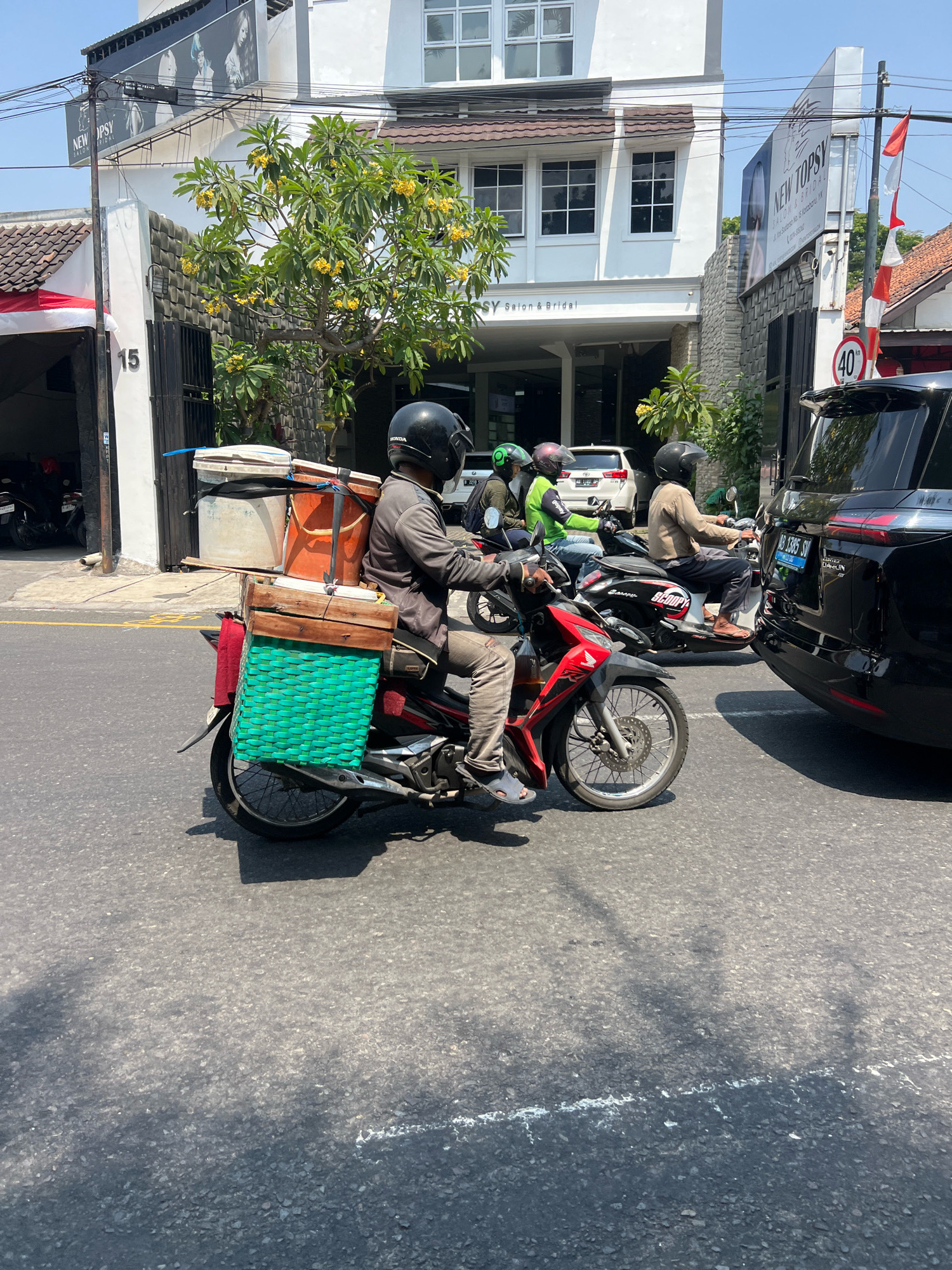 A motorcycle rider, wearing a dark helmet and grey jacket, transports several large containers and a green woven basket on the back of his red and black motorcycle. He is in the middle of a street in Yogyakarta, Indonesia, with a black SUV and other motorcycles in the background. The buildings lining the street are modern, with white facades and large windows, one of which is a salon named "NEWTOPSY Salon & Bridal". The sky is clear and blue, indicating a sunny day.