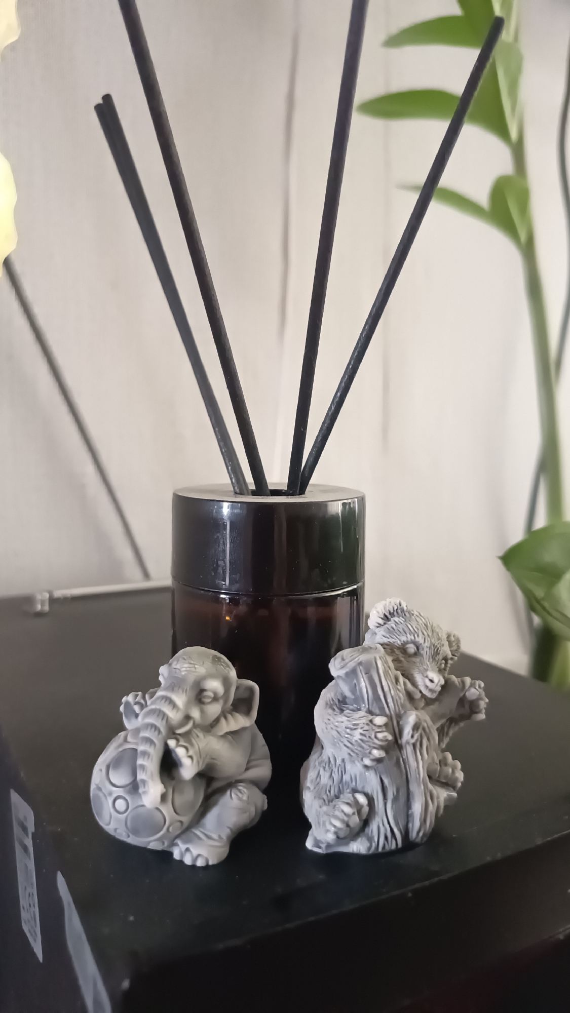 A close-up shot shows a reed diffuser with a dark brown bottle and several black reeds. To the left of the diffuser, a small, grey, elephant-shaped figurine is depicted, holding a round object with circular indentations. To the right of the elephant is a grey, bear-shaped figurine, also small, appearing to be seated. Both figurines are detailed and appear to be made of a stone-like material. The background is a lightly textured, neutral-colored surface, possibly a wall, with a plant visible on the right side, out of focus. The scene is set on a dark, flat surface.