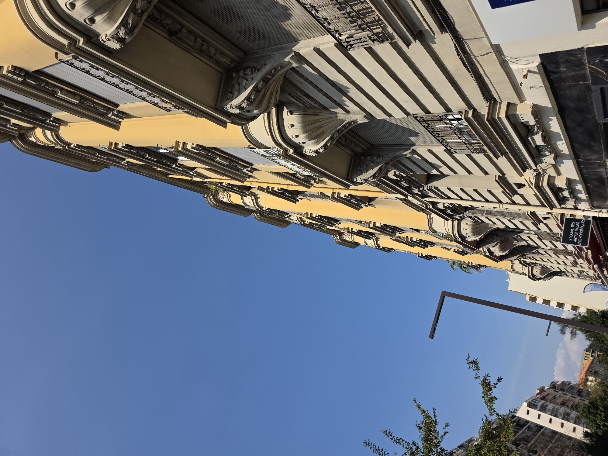 The image, taken on a clear, sunny day in Nice, France, captures an upward view of an ornate, yellow-hued building with a clear blue sky as the backdrop. The building features classical architectural elements, including decorative balconies with wrought-iron railings supported by elaborate stone brackets, and horizontal decorative friezes. The windows appear to have gray rolling shutters. To the left of the main building, other apartment buildings with balconies and trees can be seen. A modern, dark gray street lamp with a distinctive angular design stands prominently in the mid-ground. A small portion of a sign with French text, including "SPECIALITÉS ARTISANALES GASTRONOMIQUES", is visible at the bottom right of the frame, indicating a food-related establishment. The perspective is from street level, looking upwards at an angle, emphasizing the scale and architectural detail of the building.