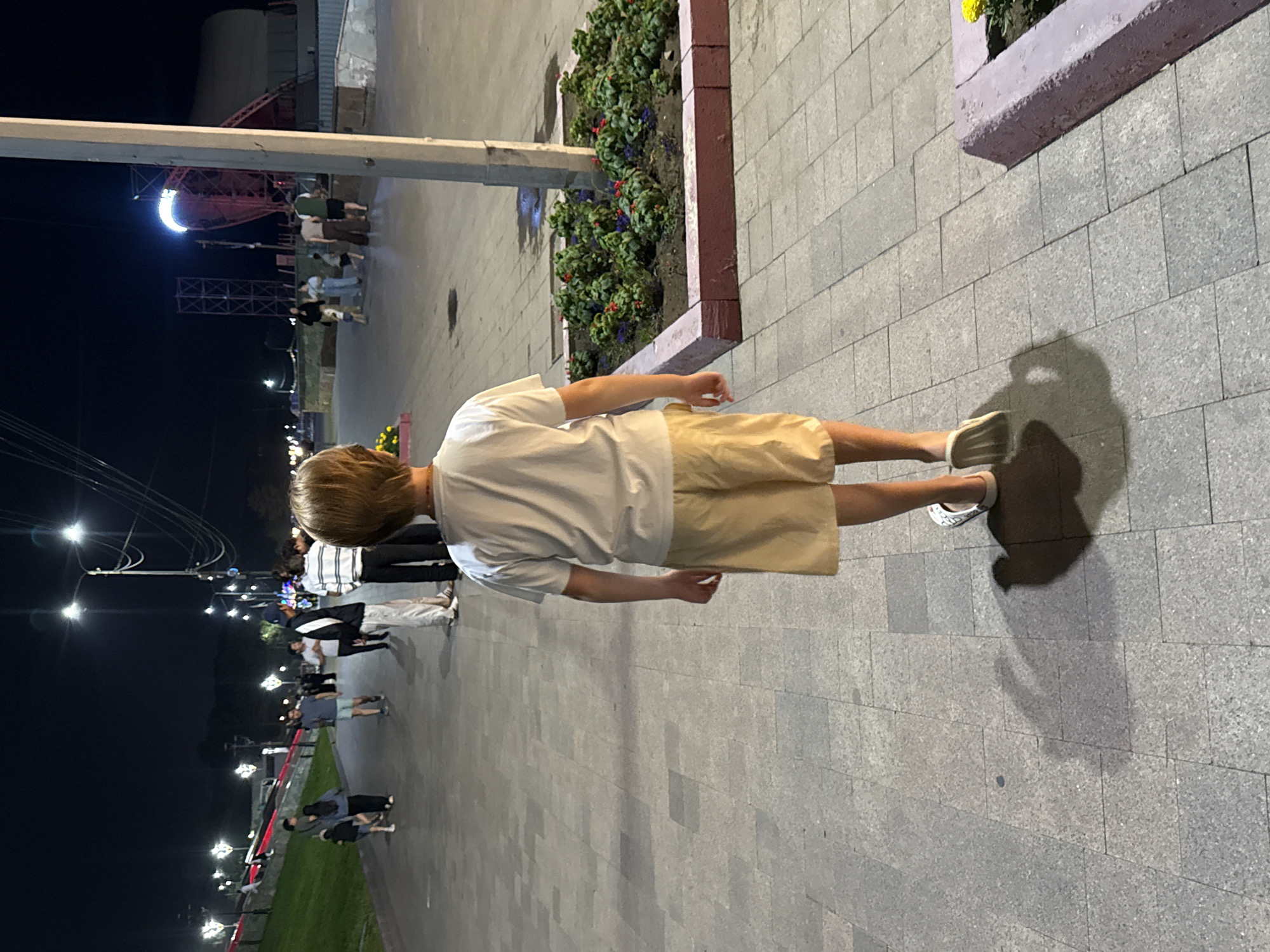 The image captures a nighttime scene on a paved walkway in Pavlodar, Kazakhstan. In the foreground, a young person wearing a white t-shirt and tan shorts walks away from the camera, casting a shadow on the ground. The walkway is made of light-colored square pavers. Behind the person, other individuals are visible walking on the path and on a grassy area to the left. Streetlights illuminate the scene, casting a warm glow. In the background, there are glimpses of buildings and structures, including what appears to be an amusement park ride. The environment suggests a public outdoor space, possibly a park or promenade, at night.