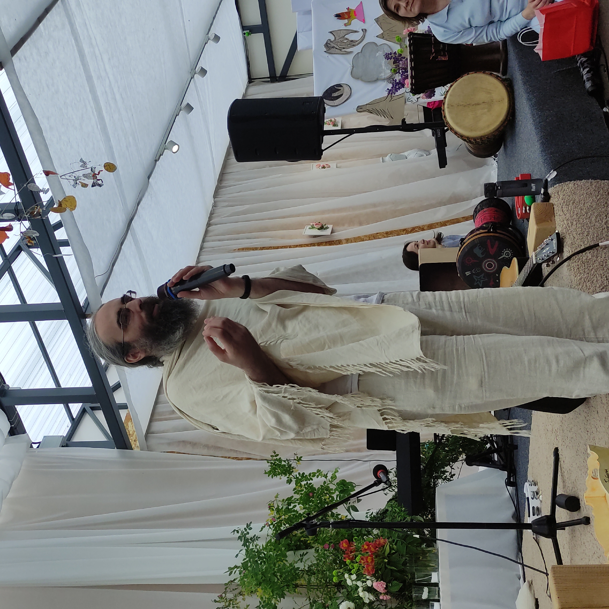A man with a beard and glasses, wearing a cream-colored robe and pants, is speaking into a microphone. He stands on a stage or platform decorated with white drapes. In front of him, there are various musical instruments including drums, a guitar, and a tambourine. Behind him, a woman in a blue sweater is seated, and further back, there are more drapes and decorative elements on the wall. The scene appears to be an outdoor event, possibly a cultural gathering or performance. The location is identified as Zhirnosovo, Russia.