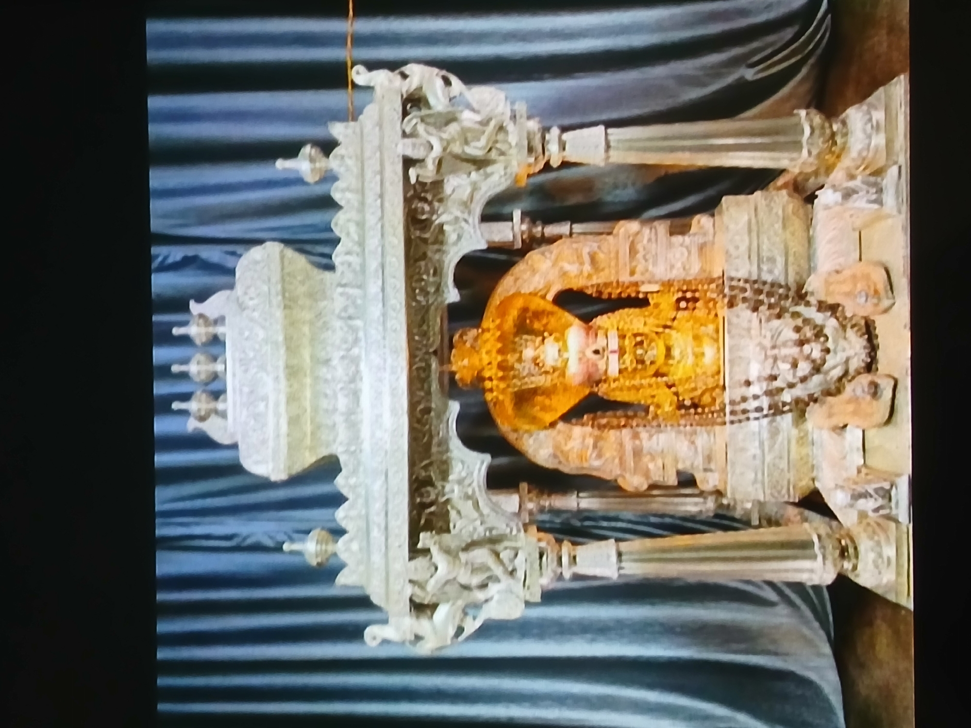 The image shows a statue of a deity, possibly a Jain Tirthankara, encased in an ornate silver-colored structure with gold accents, likely located in a temple or shrine. The backdrop appears to be draped fabric, possibly velvet, in shades of blue. The statue is richly adorned with jewelry and possibly robes. The silver structure surrounding the statue is highly detailed with decorative elements. Based on the architectural style and religious iconography, it is possible this image is from a Jain temple in Bengaluru, India, although specific identification requires further information.