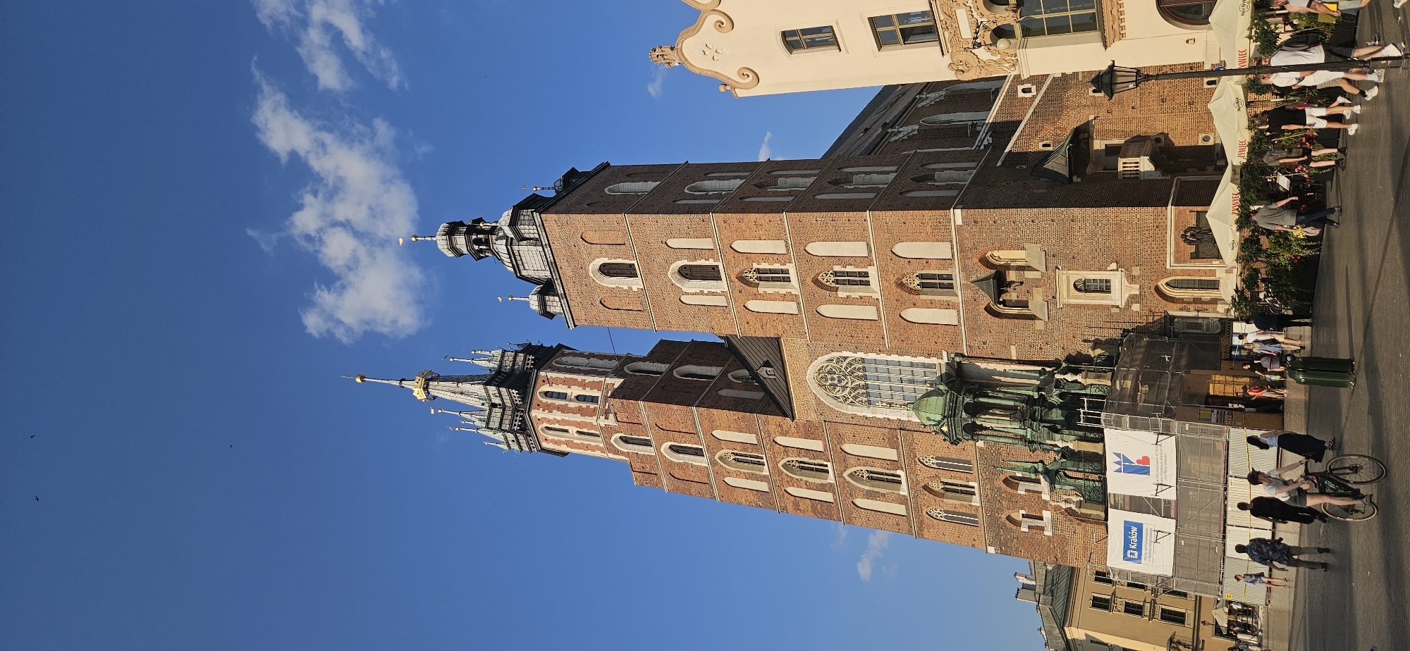 The image captures a daytime view of St. Mary's Basilica in Krakow, Poland, with its distinctive brick towers against a clear blue sky. The basilica's facade features Gothic architectural elements, including pointed arches and a large rose window. Scaffolding and banners are present on the lower section of the basilica, suggesting ongoing renovation or maintenance work.
In the foreground, the Market Square is bustling with activity. Several individuals are walking, with one person riding a bicycle. People are gathered around outdoor seating areas of cafes, indicated by umbrellas and tables. The environment is characteristic of a European city square, with a mix of historic architecture and modern-day life. The light suggests it is late afternoon, with the sun casting shadows.