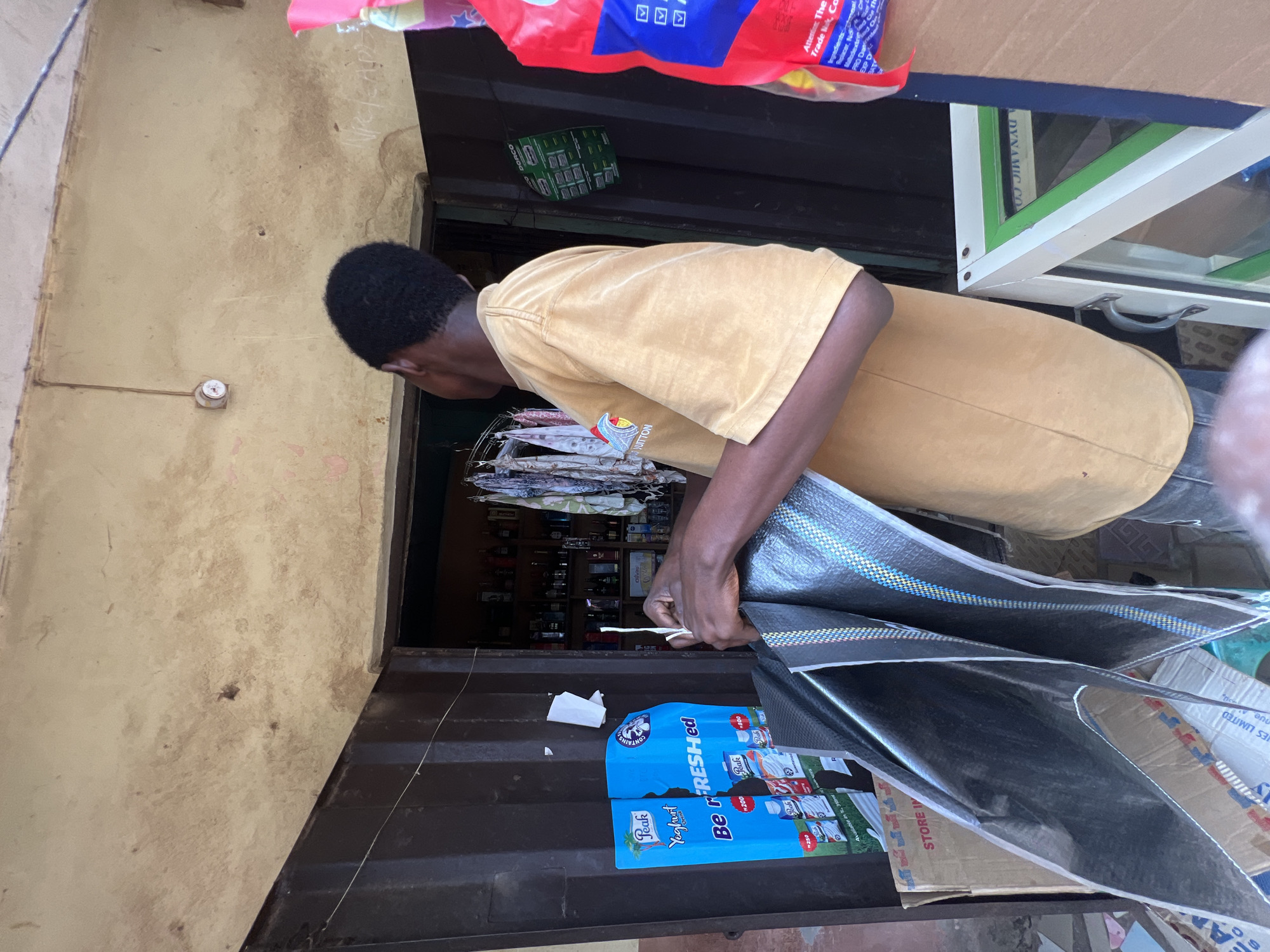 A young man, wearing a tan t-shirt with "LOUIS VUITTON" printed on it, stands in front of a small storefront in Nkpor, Nigeria. He is holding a large, folded black tarp with blue and red stripes. The storefront appears to be a small shop or kiosk with a metal door. Inside the shop, shelves are stocked with various bottles and goods. To the left of the man, a blue poster advertises "Peak Yogurt." In the foreground, a display case with a green trim is visible. The overall scene suggests a commercial setting in a developing urban area.