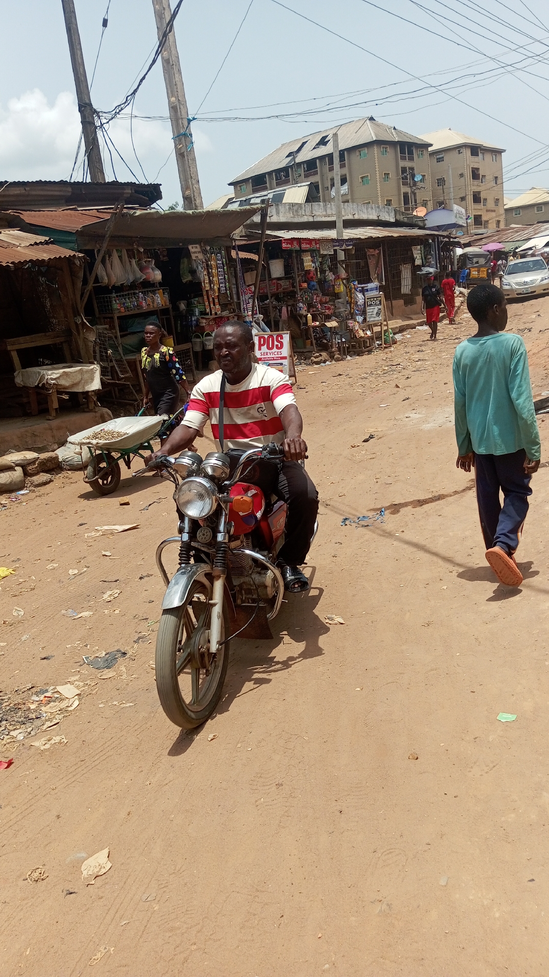 The image depicts a dusty street scene in what appears to be Onitsha, Nigeria. A man in a striped red and white shirt is riding a motorcycle on a dirt road. Stalls are lined up to the left and right of the dirt road. To the left a woman is walking with a wheel barrow filled with peanuts and to the right a boy is walking away. In the background, buildings are visible. The sky is hazy, suggesting a hot and sunny day. There is litter on the ground. A POS Service sign is present.