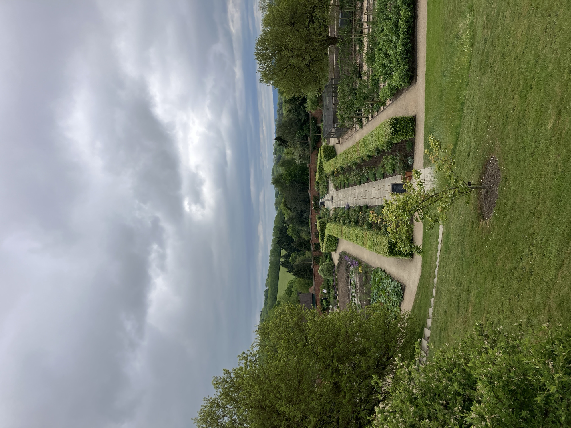 The image depicts a formal garden scene in Sevenoaks, United Kingdom, under an overcast sky. The garden features a central pathway constructed of rectangular stone blocks. This pathway is flanked by trimmed hedges and planting beds containing various plants, including what appear to be vegetables and flowers. A brick wall is visible in the middle ground, separating the formal garden from a more natural landscape of trees and fields. To the right of the formal garden, a grassy area slopes downward, featuring a young tree. The foreground includes the foliage of trees or shrubs, framing the view. The sky is predominantly grey with striations of lighter cloud cover.