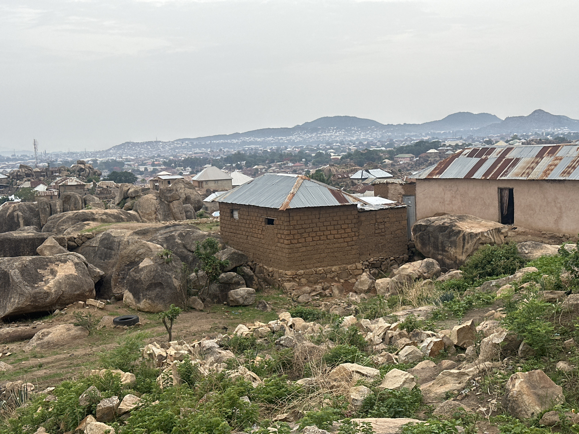 The image depicts a residential area in Jos, Nigeria, characterized by its rocky terrain and scattered buildings. In the foreground, there are large boulders and smaller rocks interspersed with sparse vegetation. Two small, single-story buildings are prominent: one made of mud bricks with a corrugated metal roof, and another with a similar roof and a door. A discarded tire rests near a smaller tree. In the background, a dense urban area extends towards a range of hills under an overcast sky. The overall scene suggests a settlement adapted to a rocky environment.