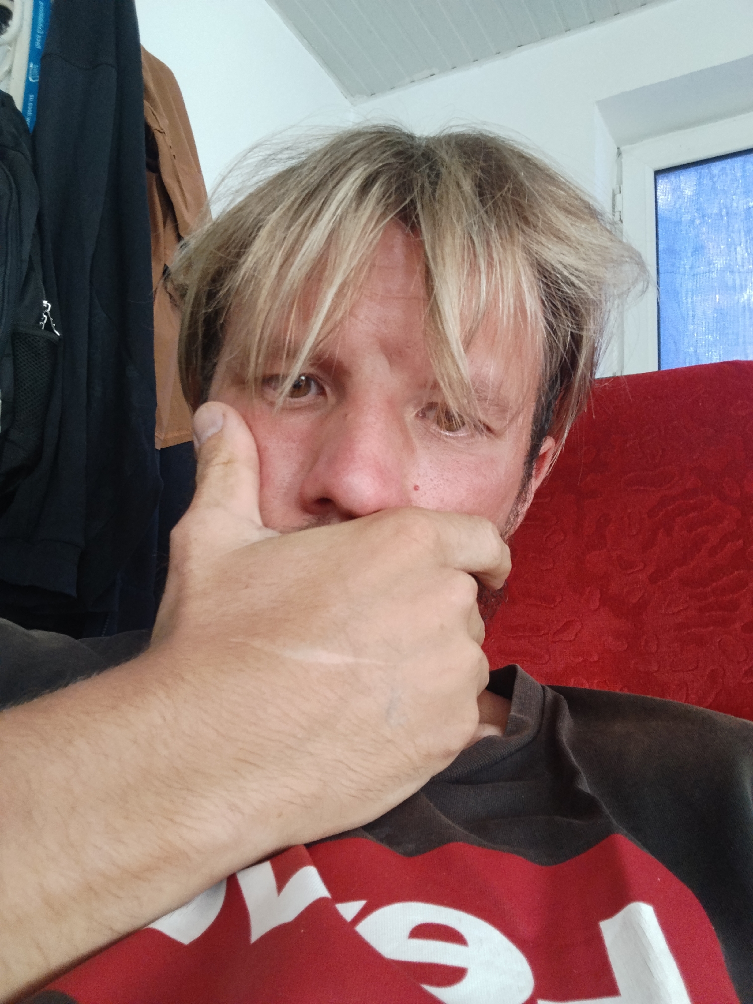 The media file shows a close-up, selfie-style shot of a person with light-colored, tousled hair and light brown eyes. The person's right hand is covering their mouth and nose, with their thumb resting against their cheek. They are wearing a dark t-shirt with a large red section and white lettering, some of which is visible. The background includes clothing hanging on what appears to be a rack or hooks, including dark fabric and a tan garment. A portion of a red upholstered chair is visible to the right, and a window with blue fabric or blinds is also present. The lighting suggests it is daytime. The setting appears to be indoors, possibly a room in a residence. No specific activity is clearly depicted beyond the person posing for the photo. The overall emotion conveyed is neutral, with the hand obscuring the lower half of the face. There is no discernible text in the visible portion of the image other than the lettering on the t-shirt, which is partially obscured. The location context of Tashkent, Uzbekistan is provided separately and cannot be inferred from the visual elements alone.