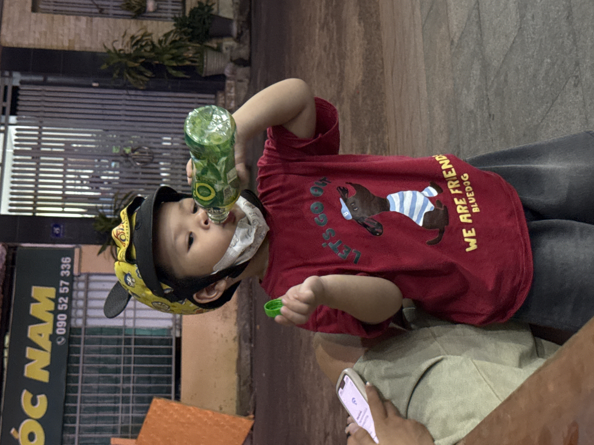 The image shows a young child wearing a yellow helmet and a red t-shirt, drinking from a green bottle. The child is also wearing a white mask. The child is holding a green bottle cap in their left hand. A cell phone is visible in the lower left corner of the image. In the background, there is a building with a sign that reads "OC NAM" and a phone number "090 52 57 336". This sign is likely for a business in Nha Trang, Vietnam. The environment appears to be an outdoor urban setting, possibly a street or sidewalk, with some plants and a metal gate visible in the background. The lighting suggests it is evening or night.
