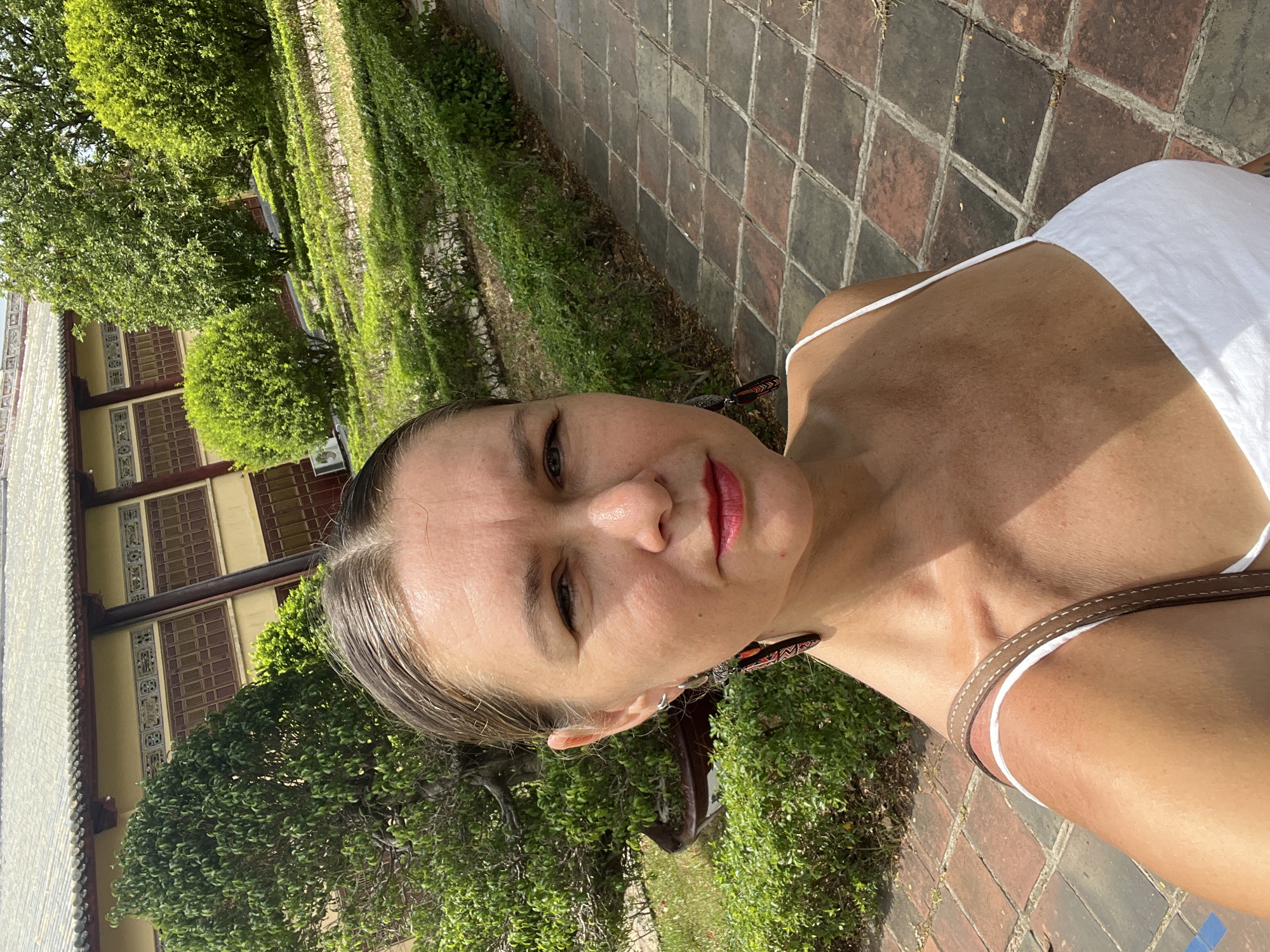 A woman stands in front of a traditional Vietnamese building with a tiled roof and yellow walls, in the city of Phú Xuân Ward, Vietnam. The building is surrounded by lush green gardens with manicured hedges and trees. The woman is wearing a white tank top and a necklace. She is looking directly at the camera, with her hair pulled back. Her expression is neutral. The foreground of the image is a stone pathway.