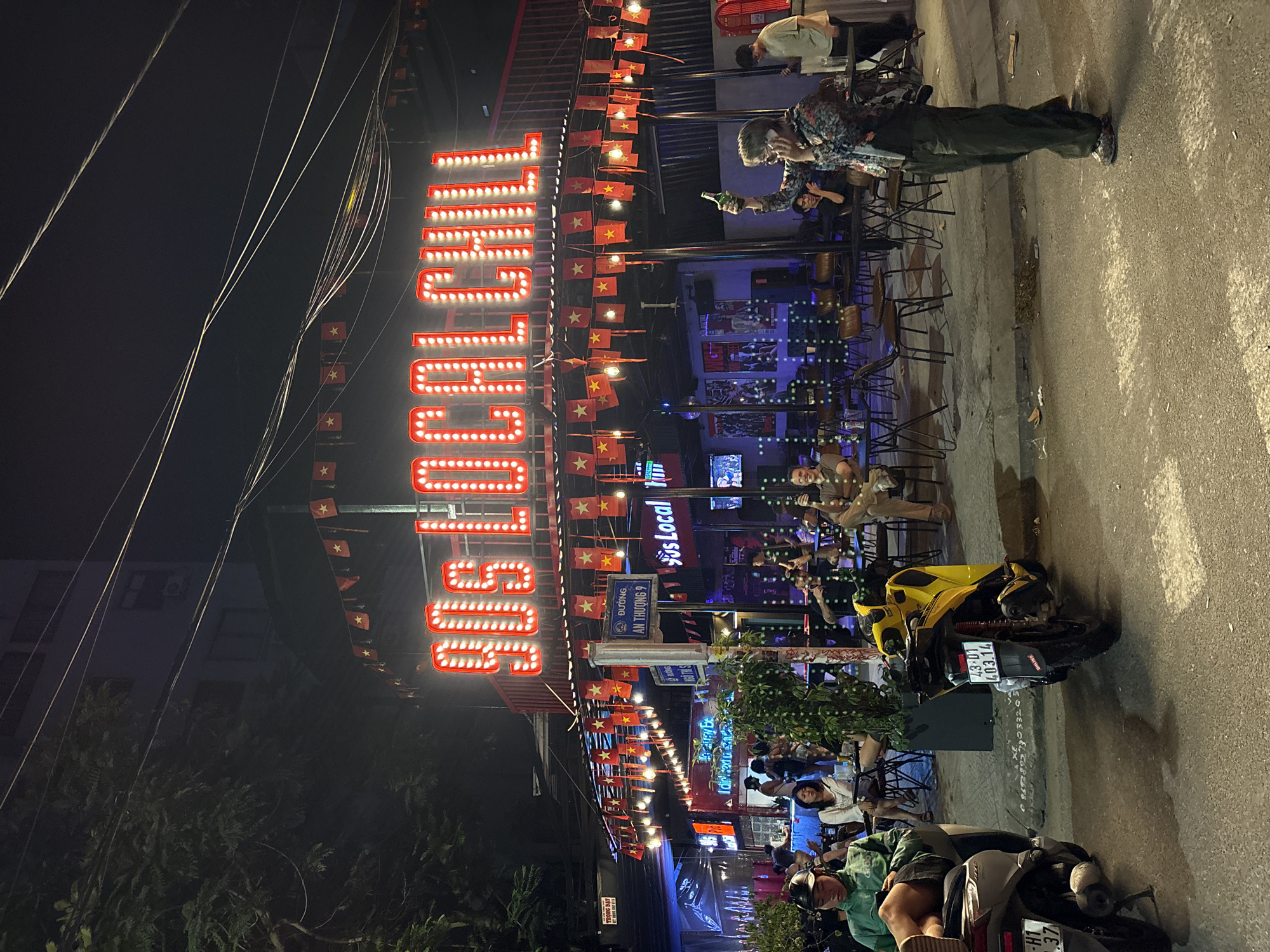 This nighttime shot captures a bustling street scene in Đà Nẵng, Vietnam, likely a commercial or entertainment district. The focal point is a bar or establishment prominently displaying the name "90S LOCAL CHILL" in large, illuminated lettering, decorated with strings of Vietnamese flags. Patrons are seated at tables, engaged in conversations and enjoying drinks. A man stands on the sidewalk, seemingly talking on his phone while holding a bottle. Several motorbikes are parked along the street, one of which is a yellow model with the license plate "43-01 403.14." The street sign indicates "ĐƯỜNG AN THƯỢNG 9." The foreground features trees and vegetation, adding to the vibrant street atmosphere. The scene is illuminated by the establishment's sign, streetlights, and other ambient lighting. Overhead power lines are visible against the night sky.