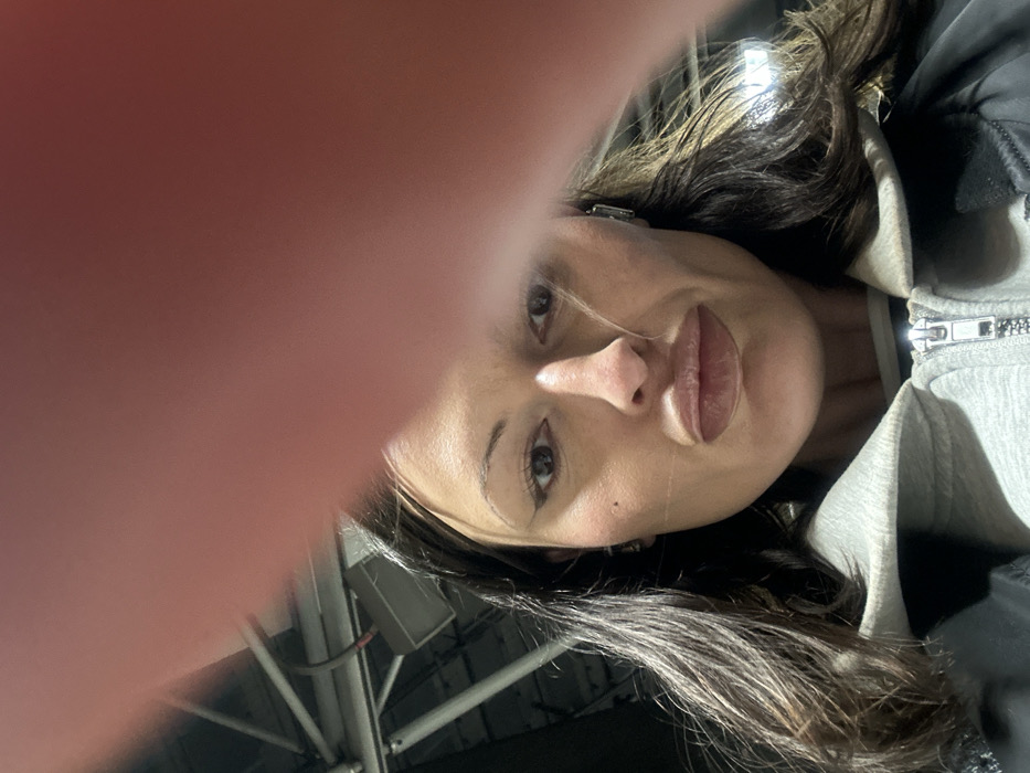 A female individual is depicted in a frontal, chest-up view, looking directly into the camera. She possesses long dark hair, winged eyeliner, and full lips. Her attire consists of a light grey hooded sweatshirt worn under a dark jacket. The upper right portion of the image is partially obscured by a large, blurred, reddish-pink object, consistent with a human finger or hand. The background is an indistinct, dimly lit indoor environment, revealing industrial overhead structures such as metallic beams and a rectangular light fixture or speaker unit. This setting is characteristic of a large public venue, like an arena or exhibition hall, located in London, United Kingdom. The specific name of the venue is not identifiable from the visual evidence.