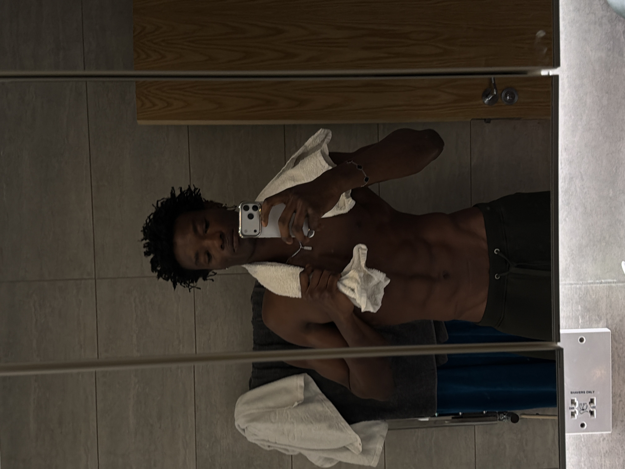 The image is a vertical, mirrored reflection of a person in what appears to be a locker room or bathroom setting, possibly in London, United Kingdom. The main subject is a shirtless individual with dark, curly hair, holding a white towel and a smartphone. The person is looking down at their phone, which is being used to capture the reflection. Their abs are clearly defined, and they are wearing dark green shorts. Another white towel is draped over their shoulder and held in their left hand.
The background of the reflection shows tiled walls and wooden shelving. A metal bar runs horizontally across the image, likely part of the mirror or its frame. On the right side of the reflected image, a white electrical outlet is visible with the text "SHAVERS ONLY" printed on it, along with various symbols and voltage indications (e.g., "230V").
The lighting suggests an indoor environment, possibly artificial, and there are no clear indicators of the time of day or weather. The overall mood conveyed is casual and perhaps post-workout, given the towels and the athletic physique. The reflection is slightly distorted by the nature of mirrors and potentially by the camera angle.