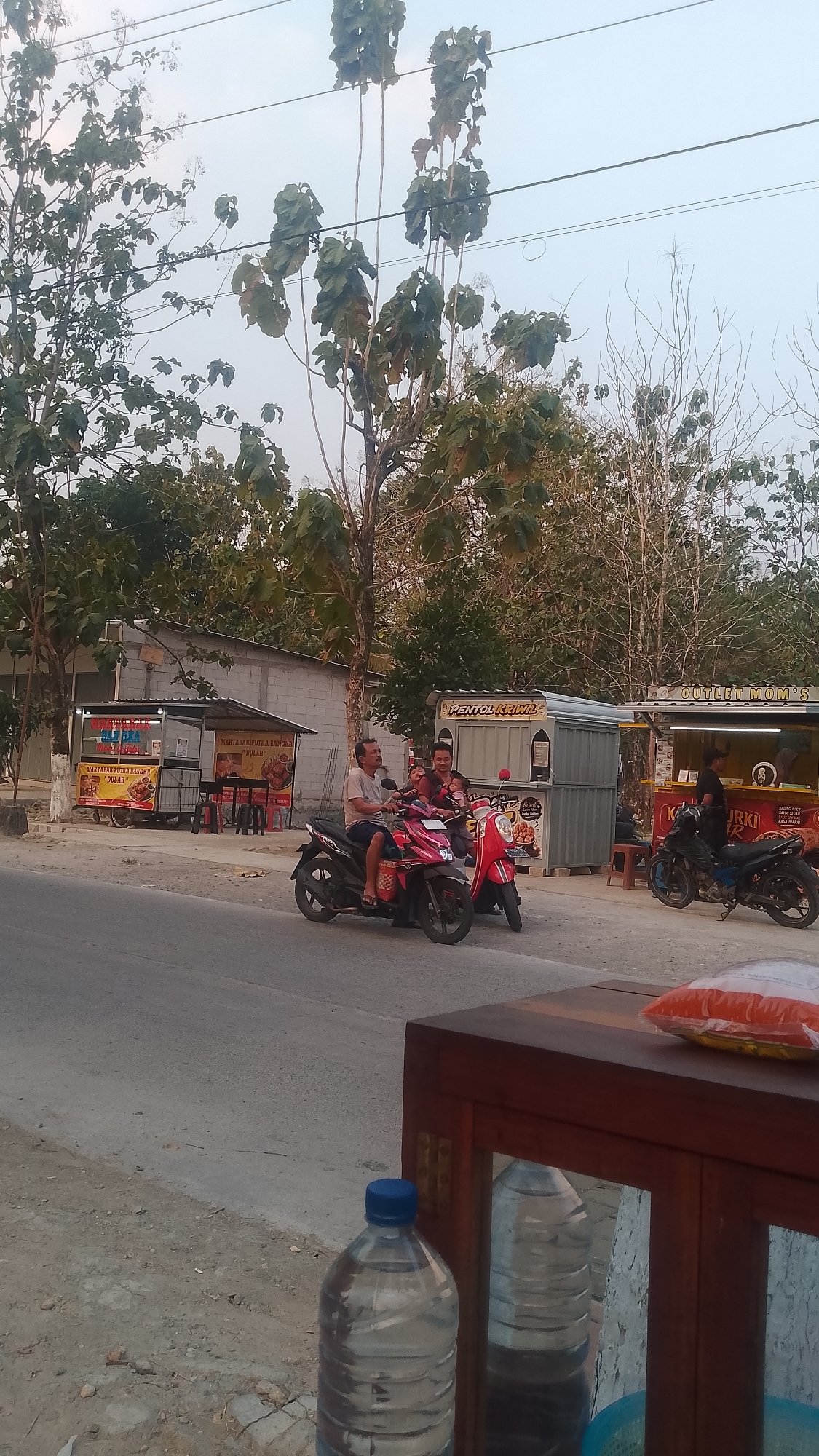 This image captures a street scene in Karanglangu, Indonesia. In the foreground, a large plastic bottle of water with a blue cap sits on a wooden surface. Behind it, a wooden cabinet with mirrored doors reflects the surroundings. To the left, a grey asphalt road runs alongside a dirt and gravel shoulder.
The middle ground features a street lined with small food stalls. On the left, a stall with a yellow and red awning displays food items. Further right, a stall with a grey roof and white walls is visible. In front of these stalls, two motorcycles are parked. A pink scooter is parked near a red and white scooter. Three people are seated on the pink scooter; a man wearing a grey shirt is driving, a woman in a blue shirt is behind him, and a child is seated in front of her.
The background is dominated by trees and vegetation. Power lines crisscross the sky above. On the right side of the frame, another motorcycle, black and blue, is parked in front of a food stall with a prominent "PENTOL KRIMIL" sign. A person in a dark shirt is visible at this stall. The overall atmosphere appears to be daytime, with a bright, overcast sky.